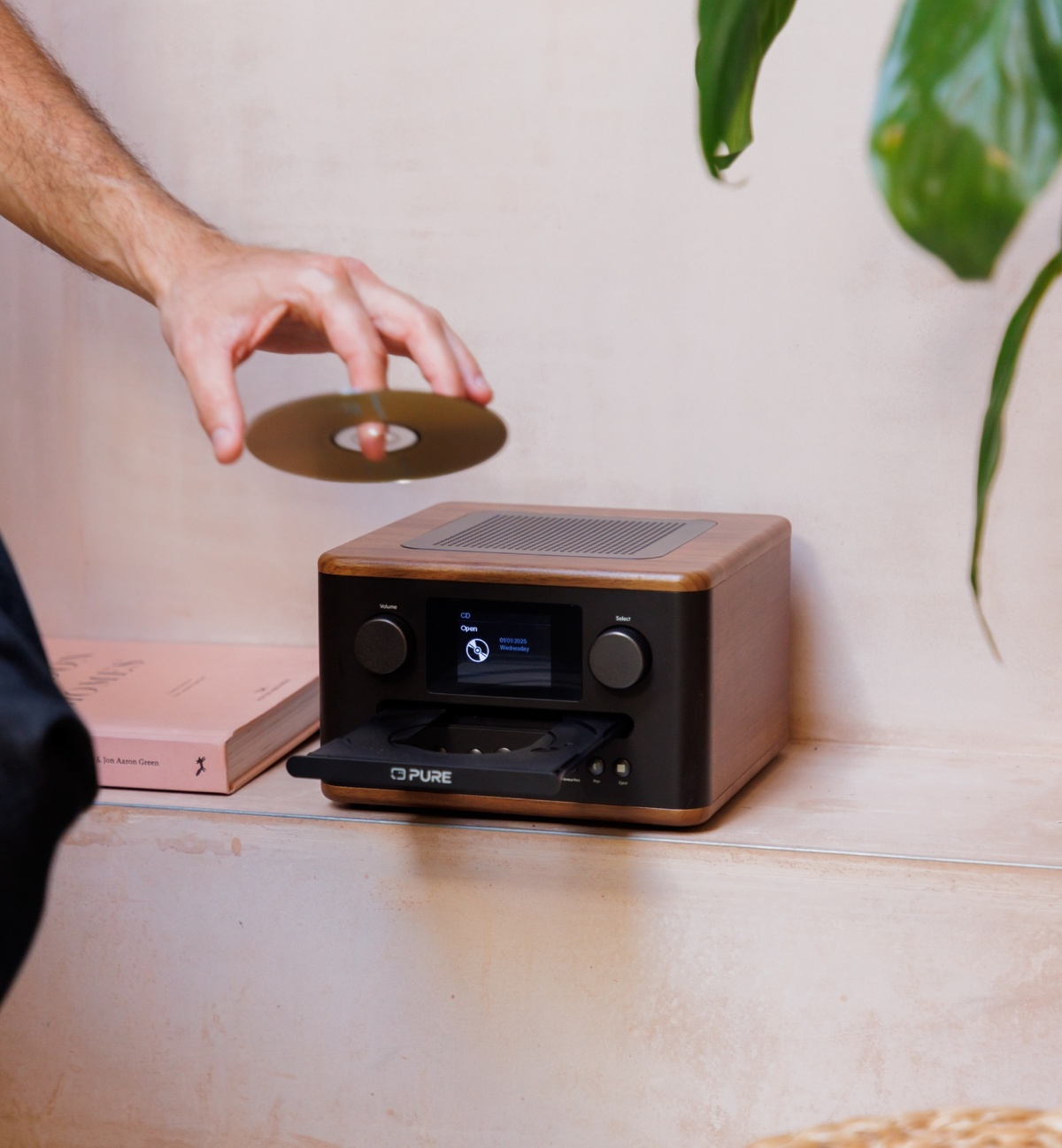 Une main place un CD dans une radio en bois. La radio a un écran affichant des informations. Un livre et une plante sont en arrière-plan.