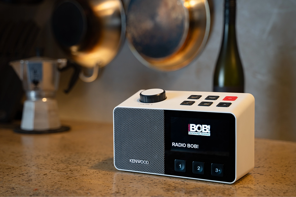 Une radio Kenwood blanche affiche 'Radio Bob !' sur l'écran. Des casseroles, une cafetière et une bouteille de vin en arrière-plan flou.