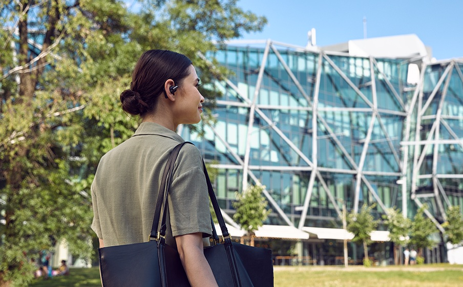 Kobieta ze słuchawkami i torbą przechodzi obok nowoczesnego budynku i drzew. Czyste błękitne niebo w tle.