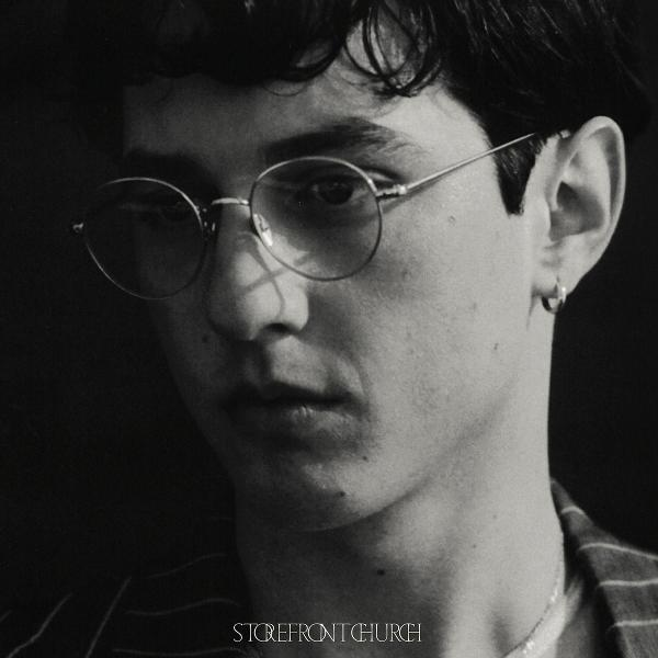 Ein Schwarz-Weiß-Porträt eines Mannes mit runder Brille, Ohrring und gestreiftem Hemd. Das Bild ist eine Nahaufnahme seines Gesichts.