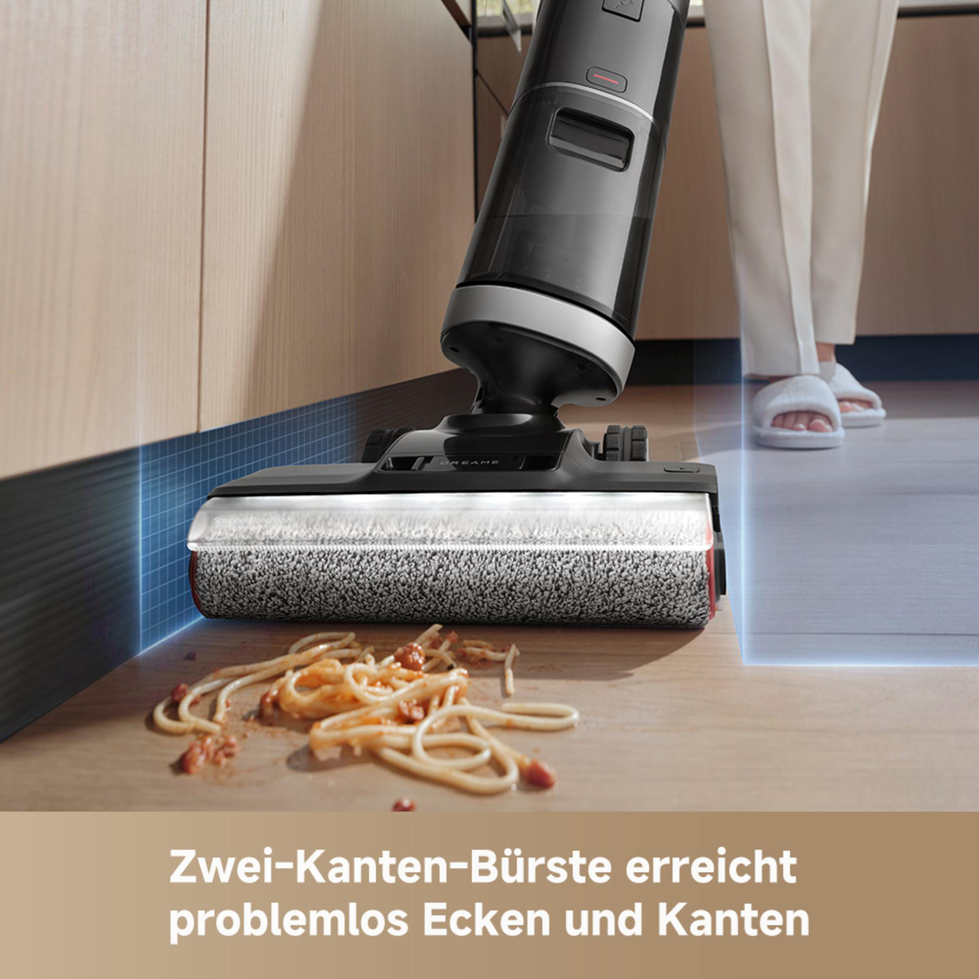 Ein Staubsauger reinigt verschüttete Spaghetti. Eine Person in weißer Hose und Hausschuhen steht daneben. Hölzerne Schränke sind im Hintergrund.