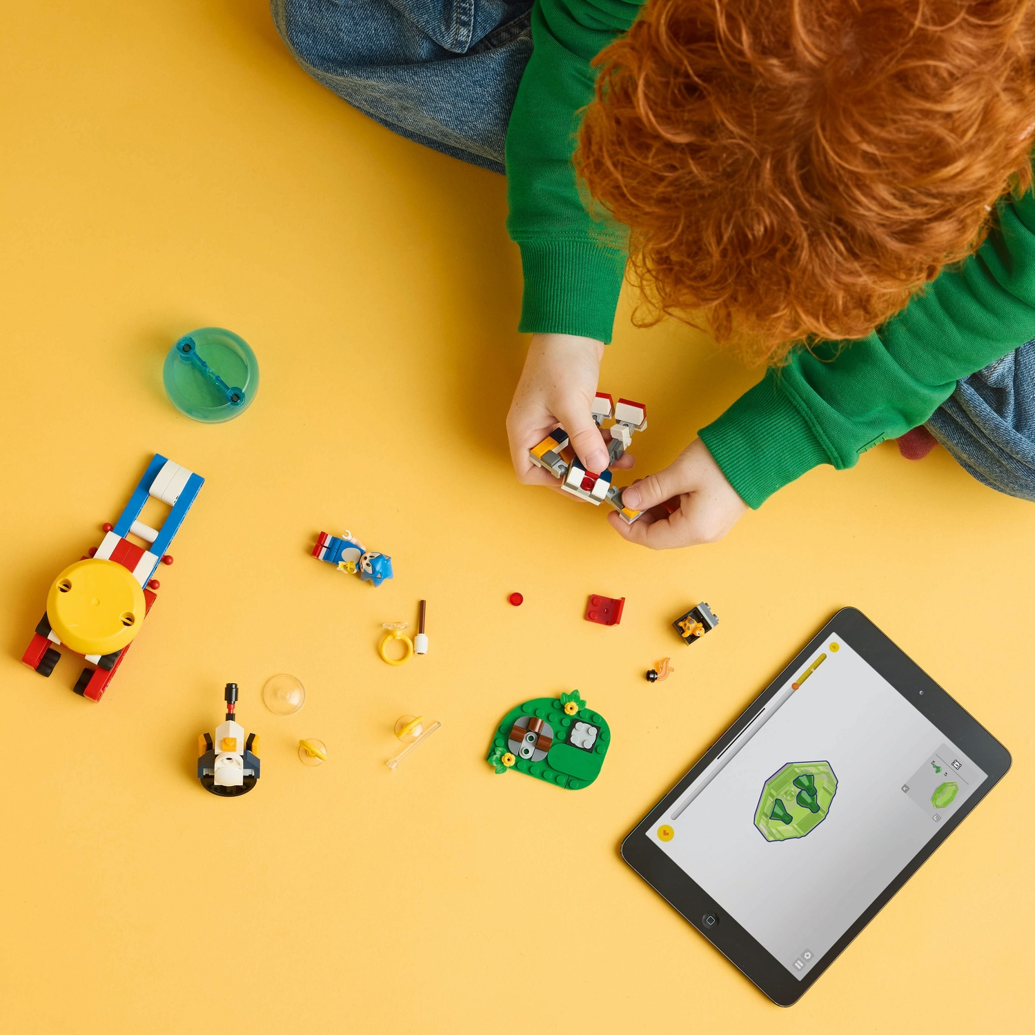 Een kind in groen speelt met Lego-stenen op een gele ondergrond. Een tablet toont een Lego-instructie. Diverse Legostukken liggen verspreid.