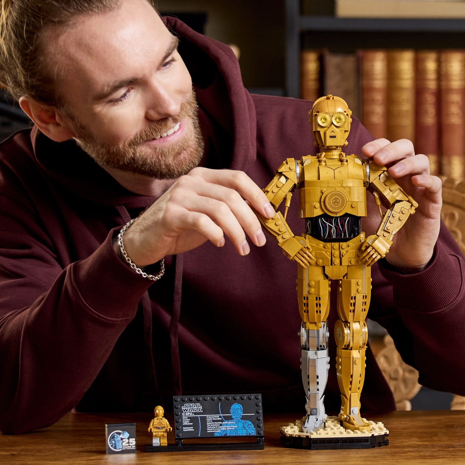 A man holds a gold Lego C-3PO figure. A small Lego C-3PO and info card sit on a table. Background has books.