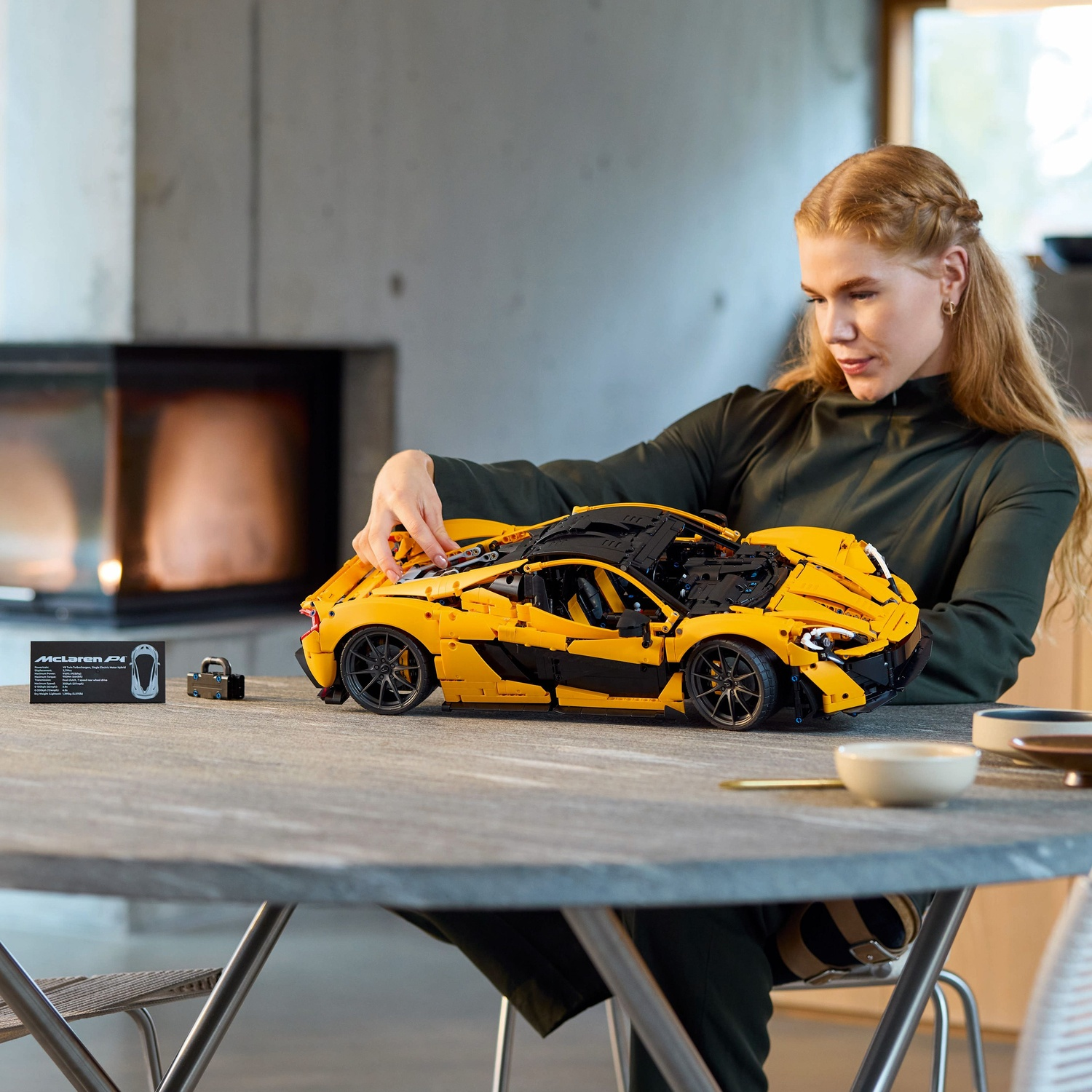 Een vrouw monteert een gele LEGO McLaren P1 op een tafel. Accessoires, waaronder een sleutel en een kaart, zijn in de buurt. Een open haard is zichtbaar.