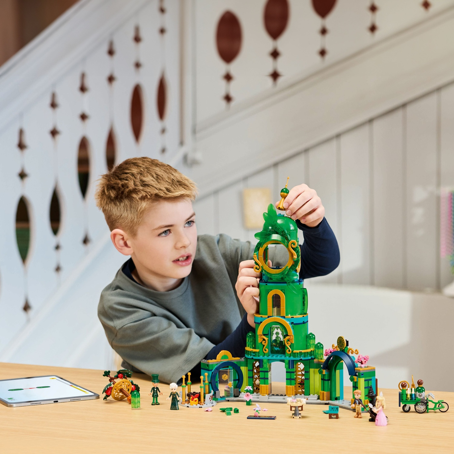 A boy builds a green Lego structure. A table holds the build and figures. A tablet sits to the side. A staircase is in the background.