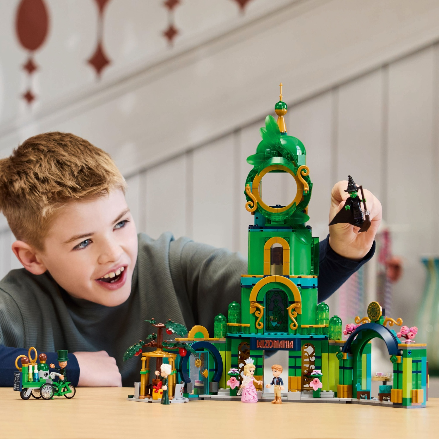A boy smiles, playing with a Lego set. A green building with 'Wizomania' is central. A witch figure is being held.