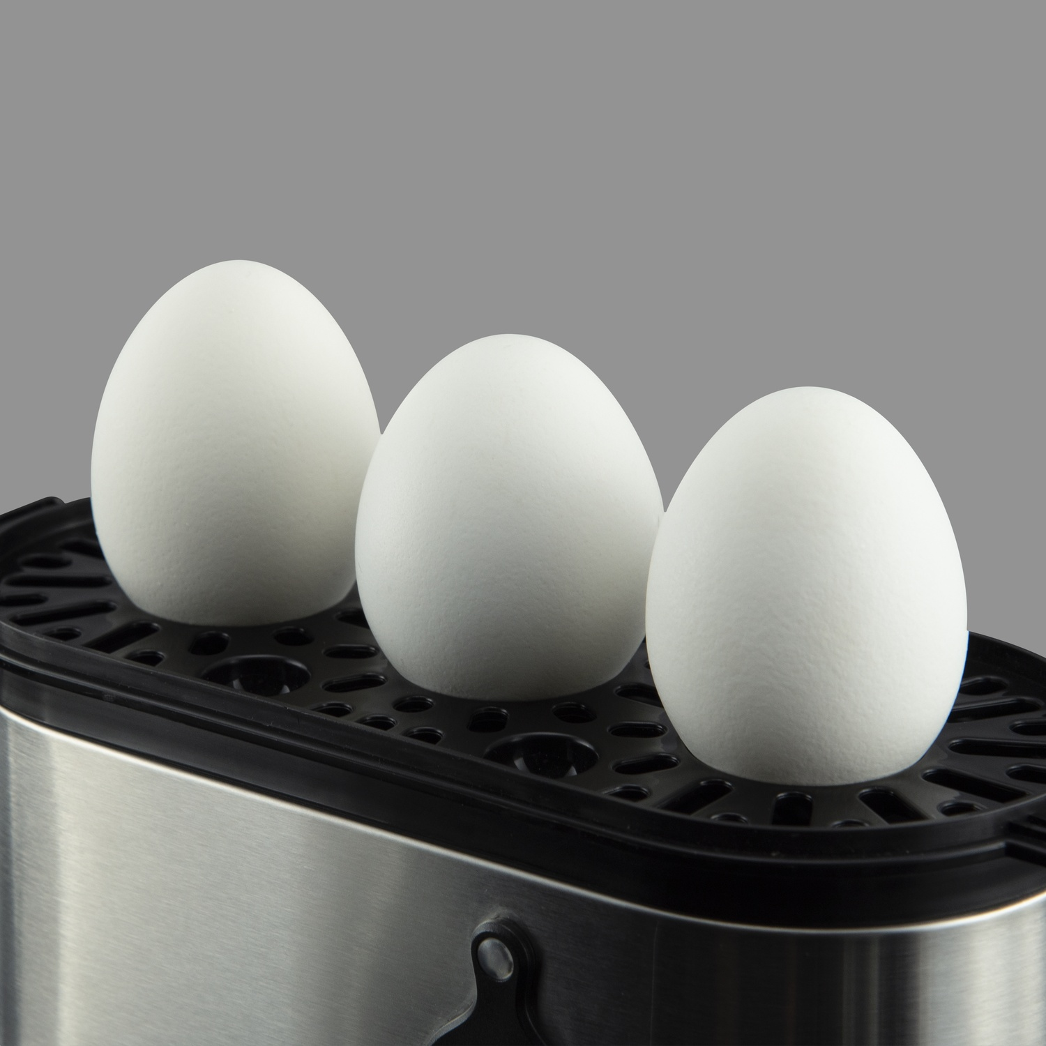 Three white eggs in an egg boiler. The egg boiler is black and stainless steel. The background is gray.