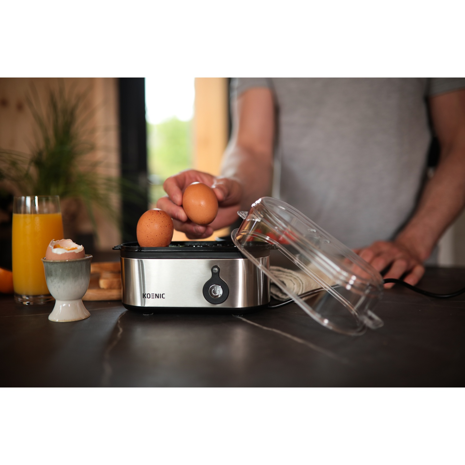 A person placing eggs in a Koenic egg boiler. There is orange juice, an egg cup with an egg, and the clear lid.