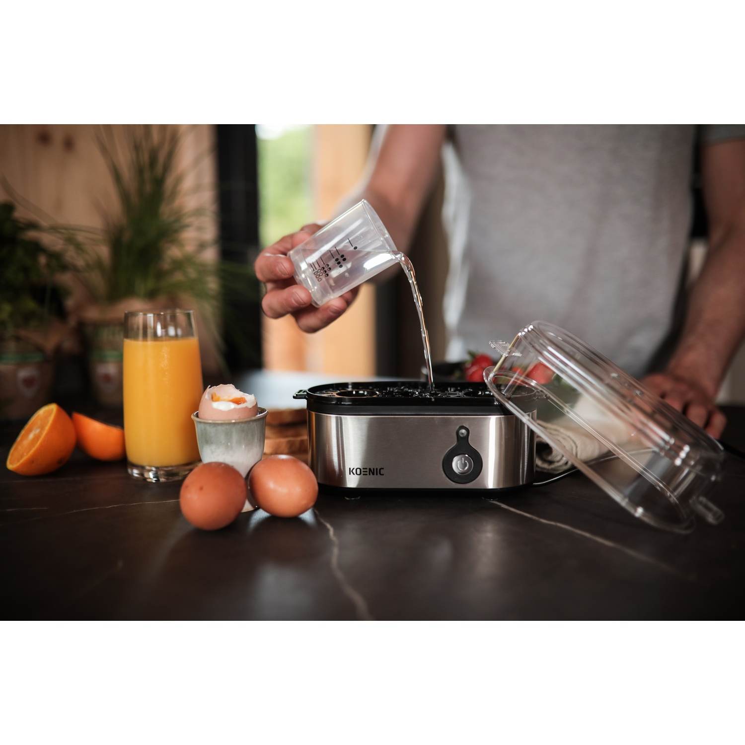 A person pouring water into an egg cooker. Eggs, orange juice, and oranges are on the table. The cooker is silver.