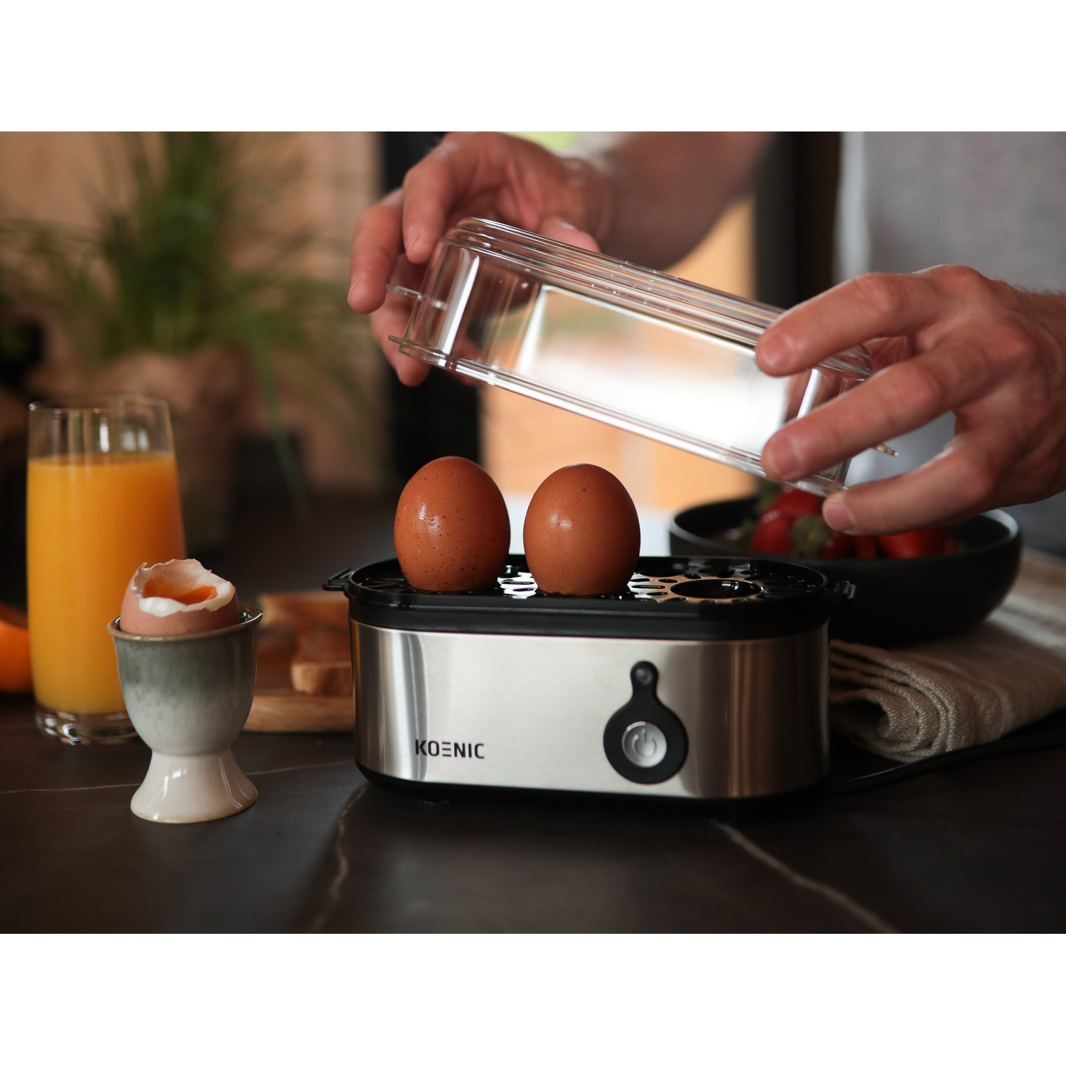 A person placing a lid on an egg boiler with two eggs. There is orange juice, a soft-boiled egg, and a bowl of strawberries.