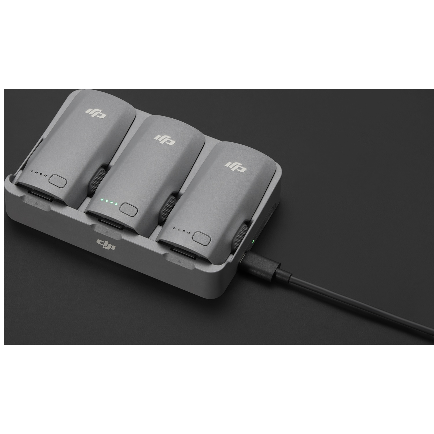 Three gray drone batteries in a charger connected with a black cable against a dark gray background.