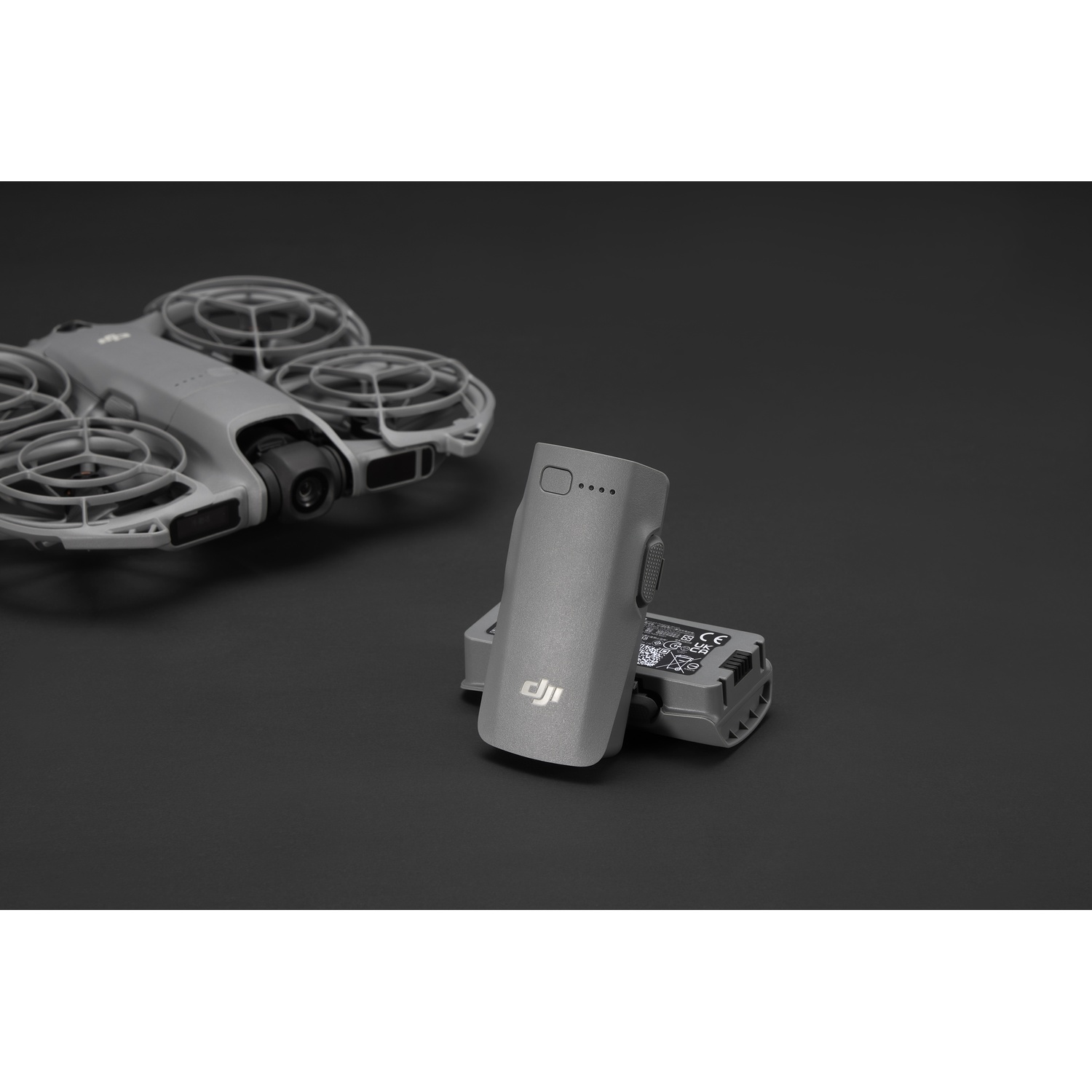 A gray drone and two gray batteries are on a black surface. One battery is upright. The drone is in the background.