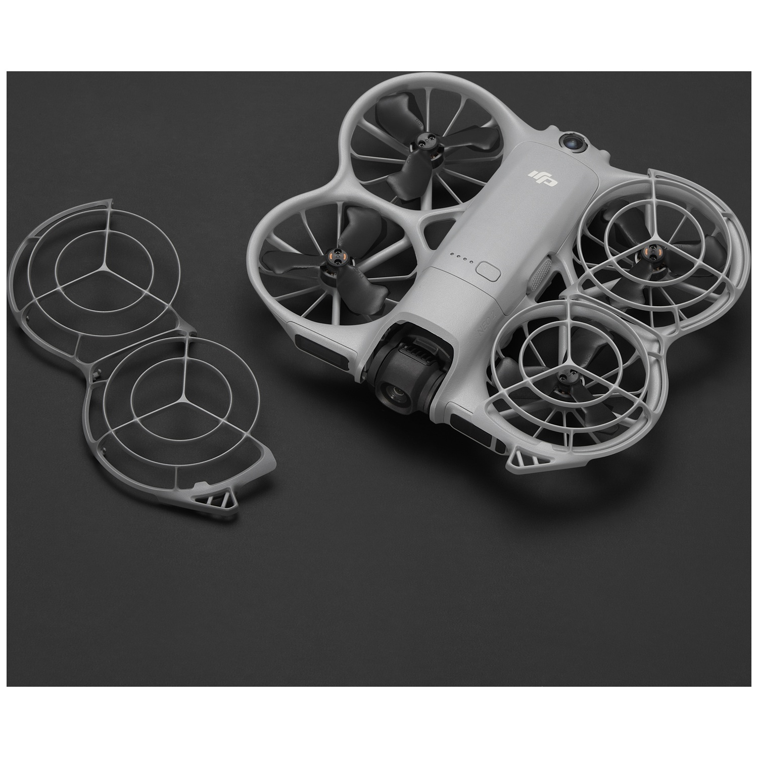 Un drone gris avec quatre hélices et un protège-hélice détaché, sur une surface noire. Le drone est incliné, révélant des détails.
