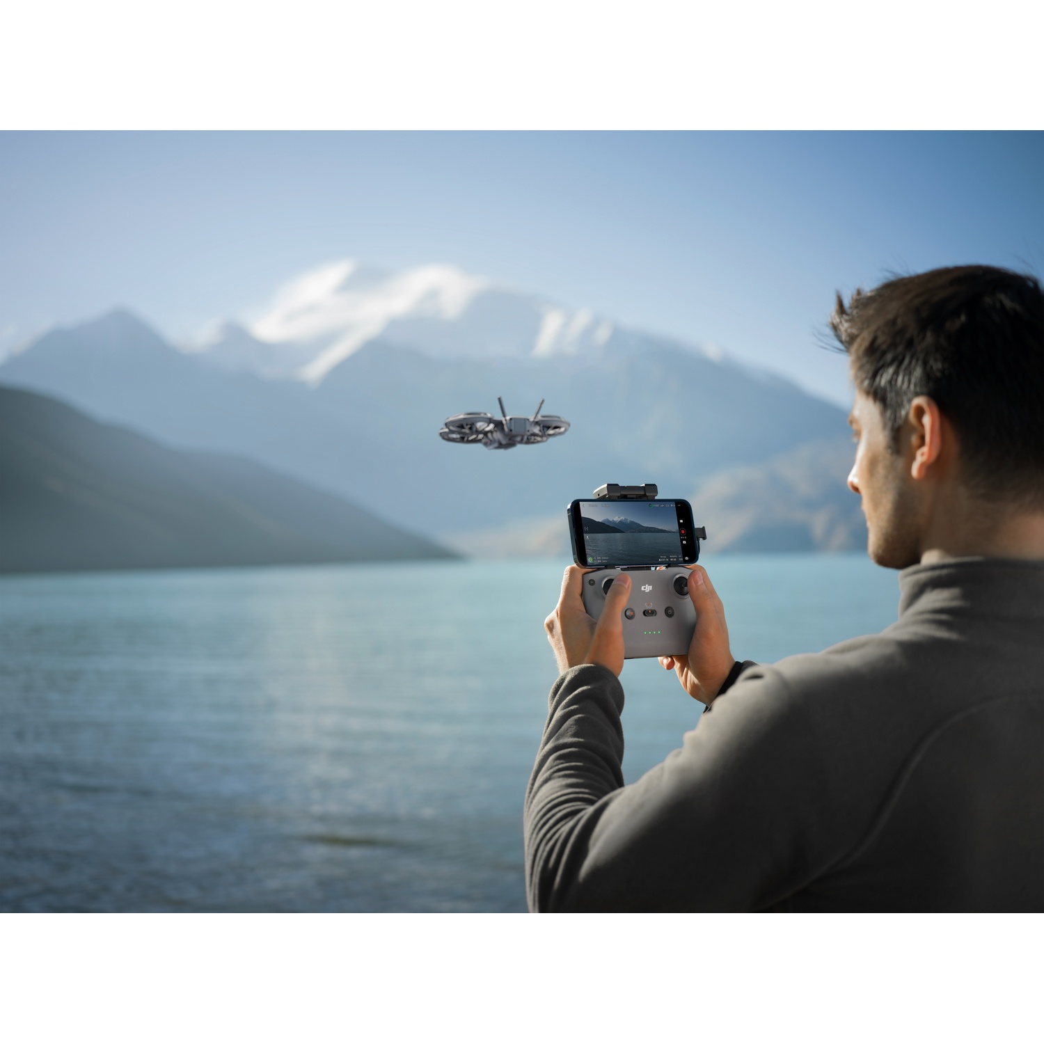 A person holding a drone controller with a phone, flying drone in the sky, lake and mountains in the background.