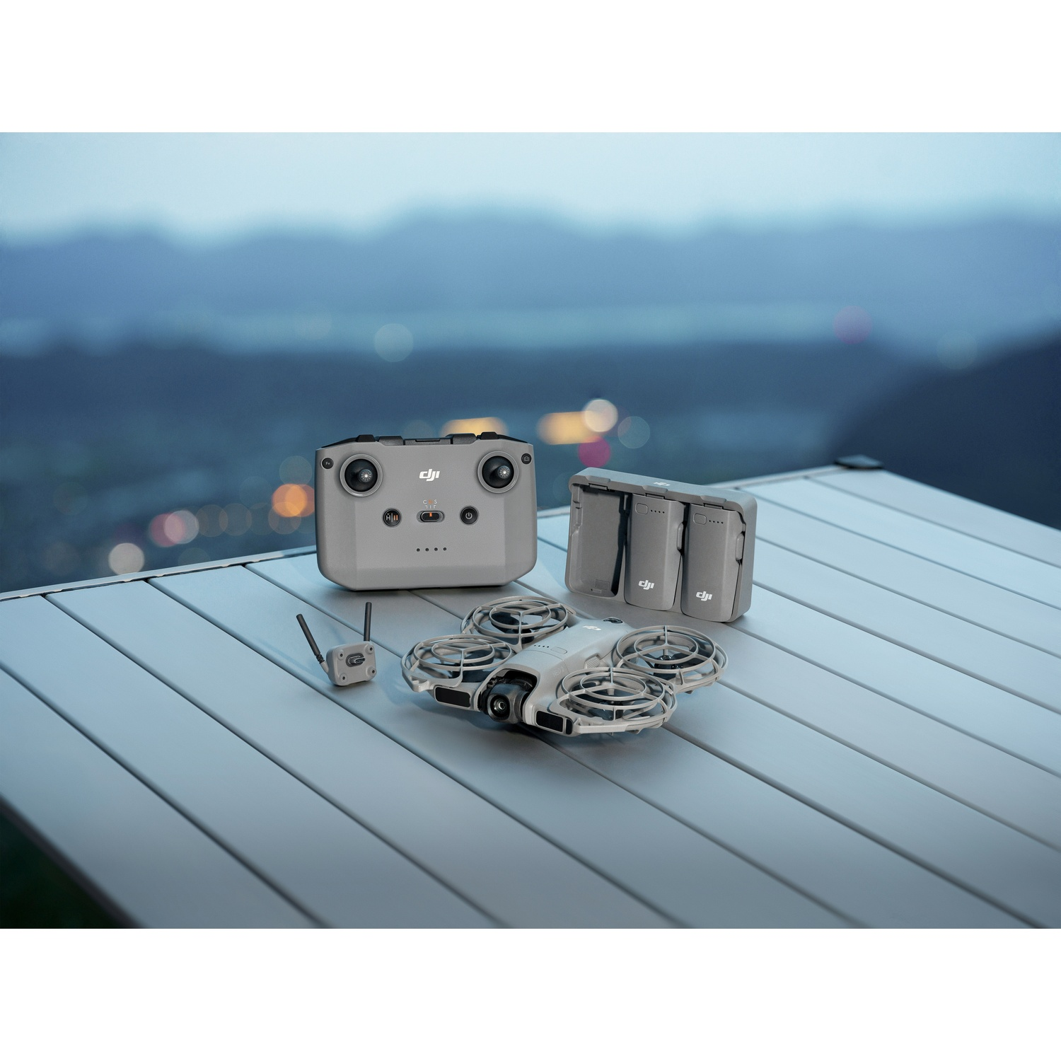 Drone, remote, battery pack on a table, with cityscape blurred in the background. Gray drone and accessories.
