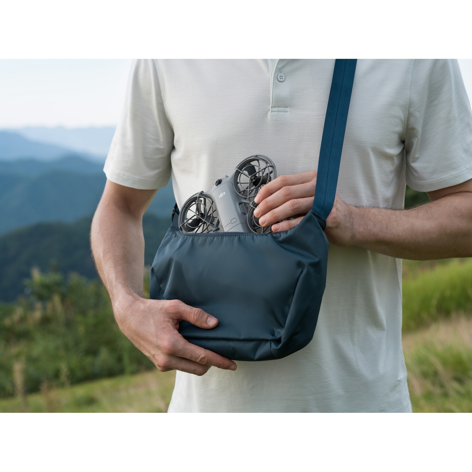 A person holding a gray bag with a drone inside. The person is wearing a light-colored shirt and is outside. Green hills are in the background.