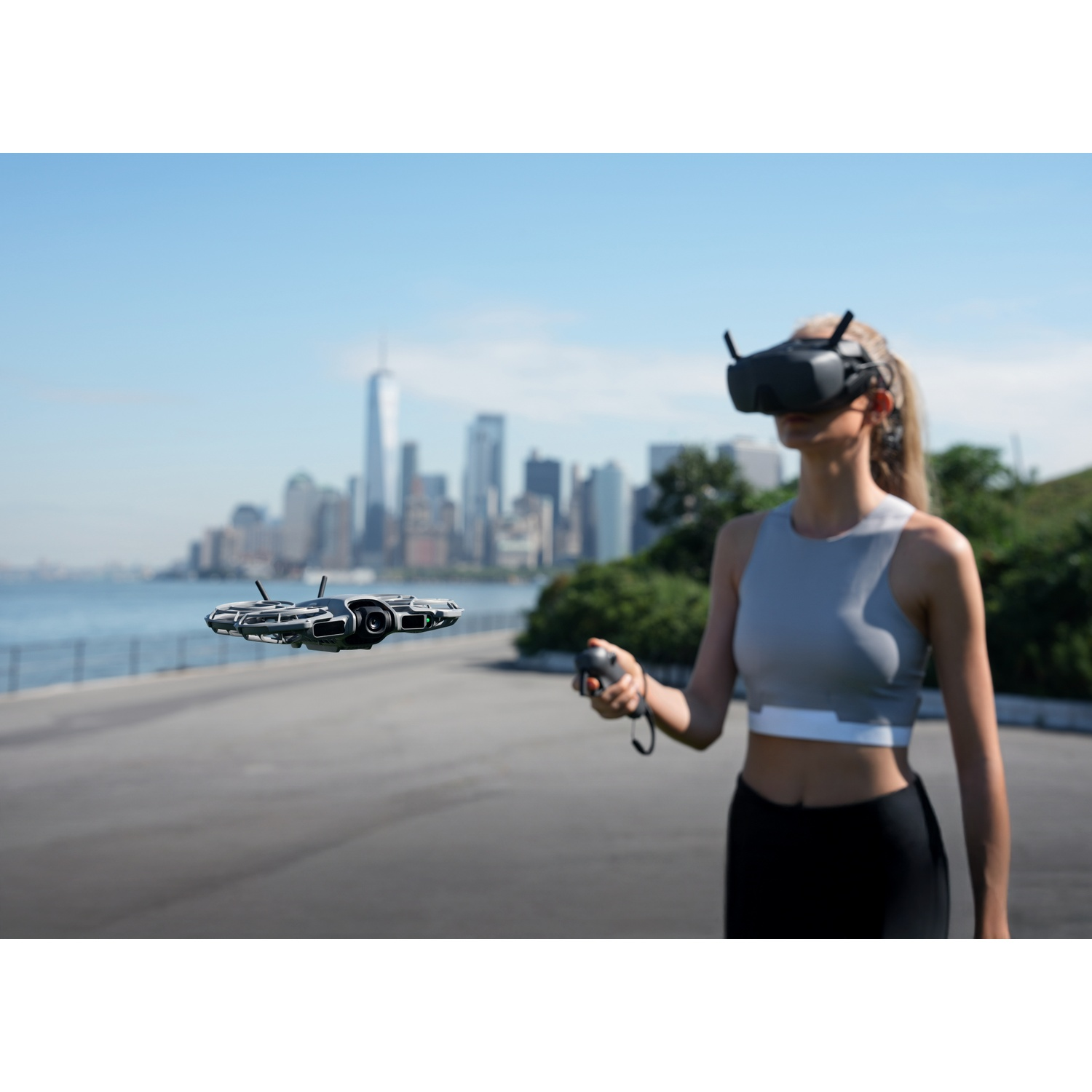A woman wears VR goggles, holding a controller. A drone hovers in front of her. A city skyline and blue sky are in the background.