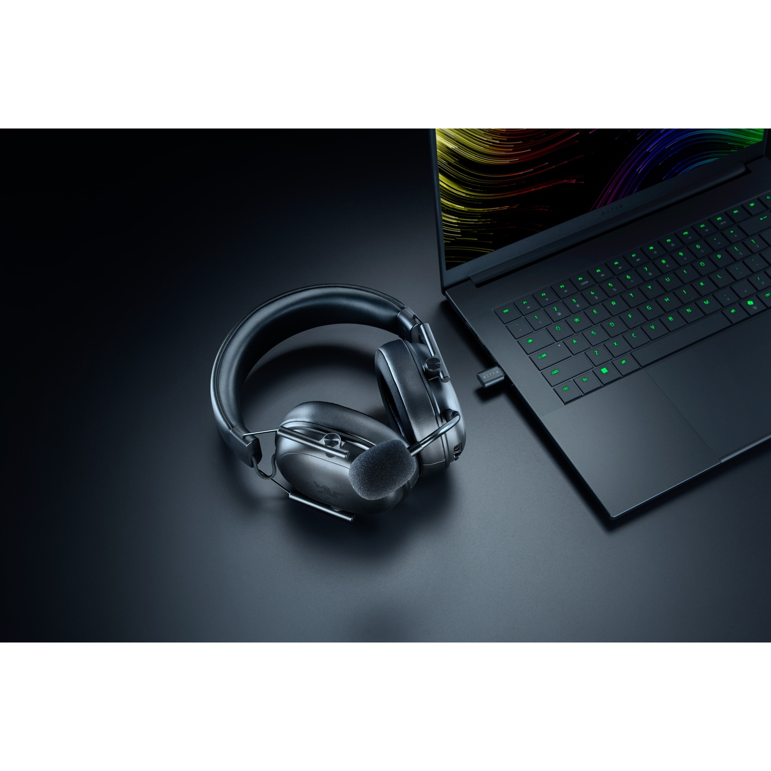 Zwarte gamingheadset met microfoon ligt naast een laptop met groen oplichtend toetsenbord, op een zwart oppervlak.