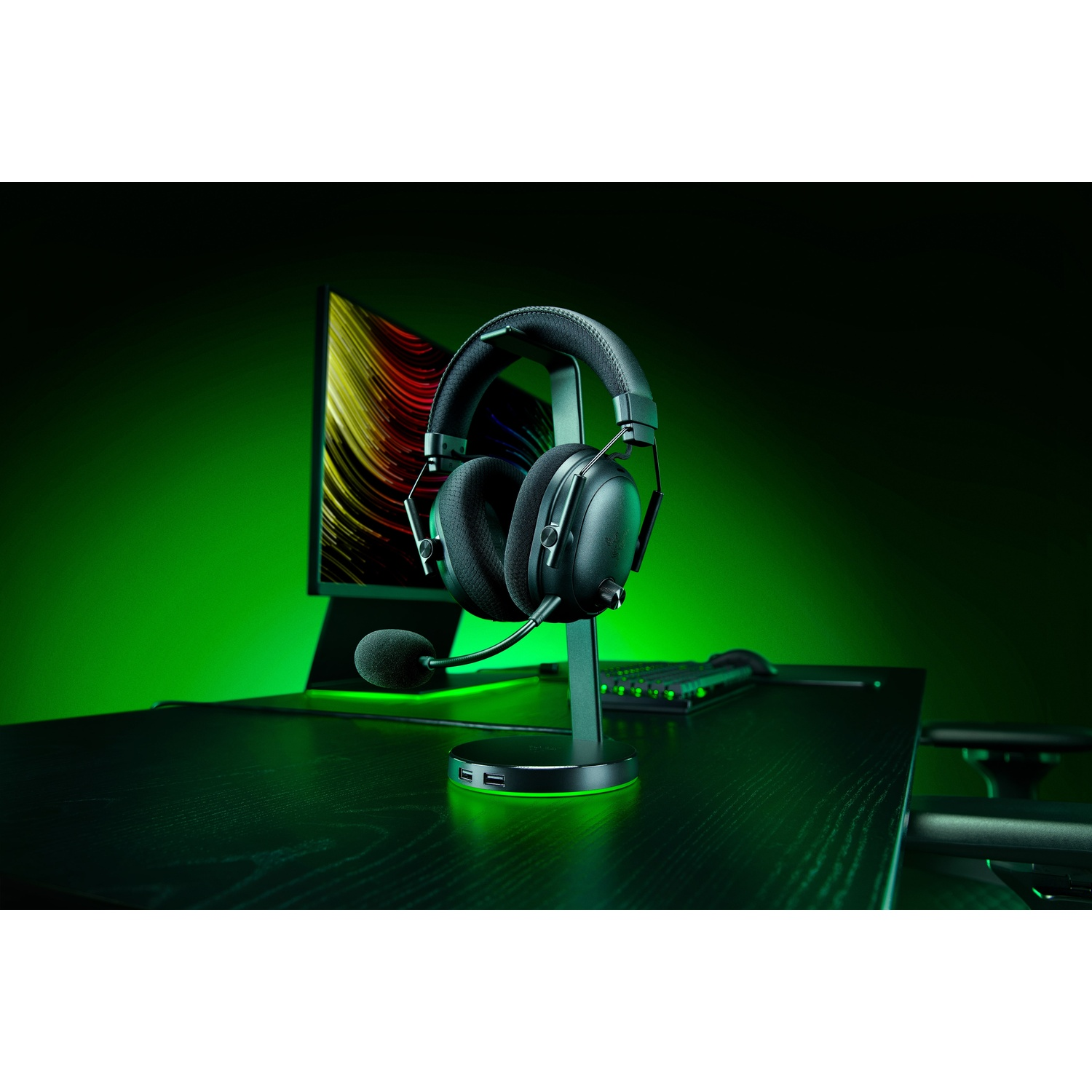 Zwarte gaming headset op een standaard, bureau met toetsenbord en monitor op de achtergrond, groene verlichting.