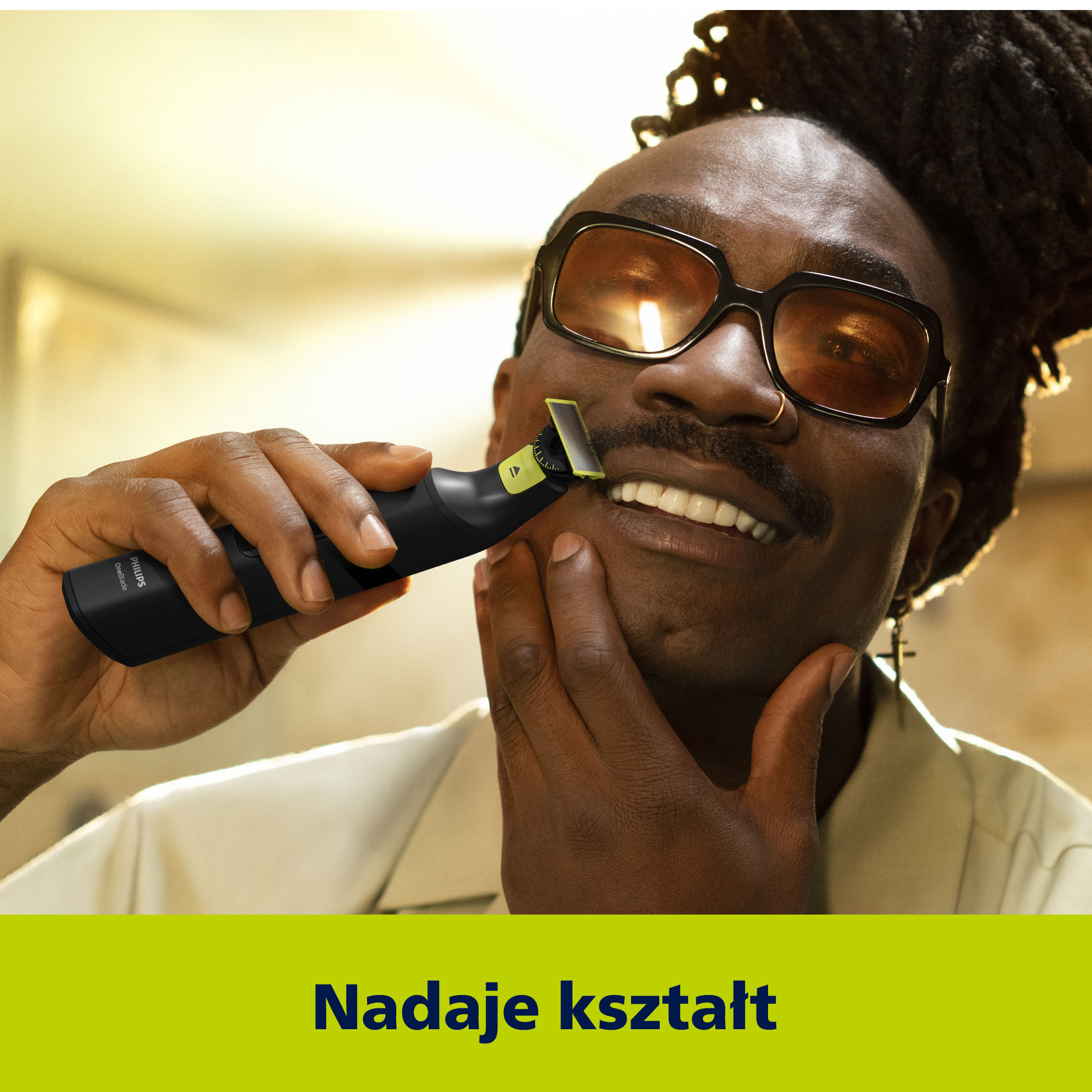 Mężczyzna w okularach goli brodę czarną maszynką. Uśmiecha się. Tło jest żółte.