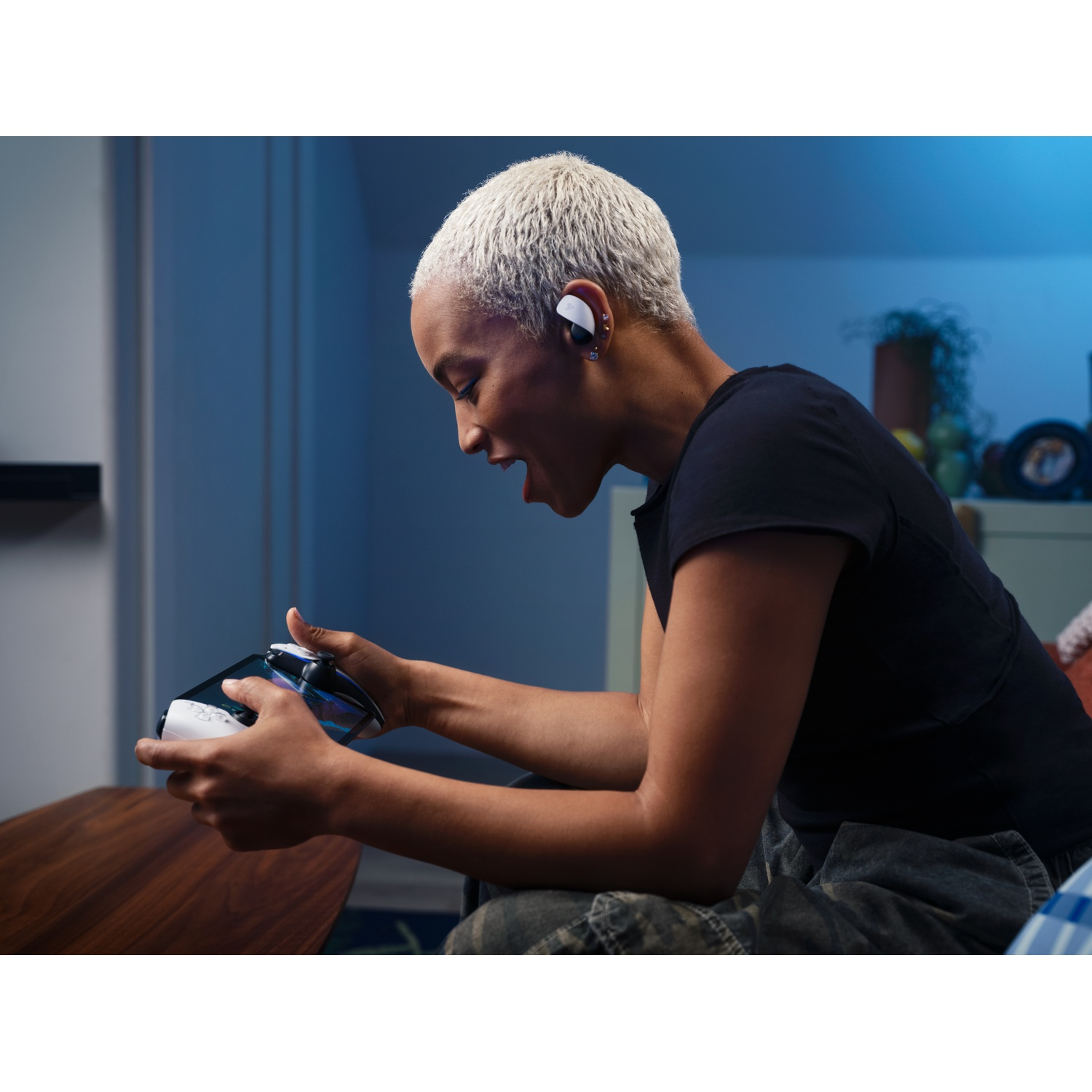 A woman with short, blonde hair plays a video game, wearing earbuds. She is holding a game controller and is excited.