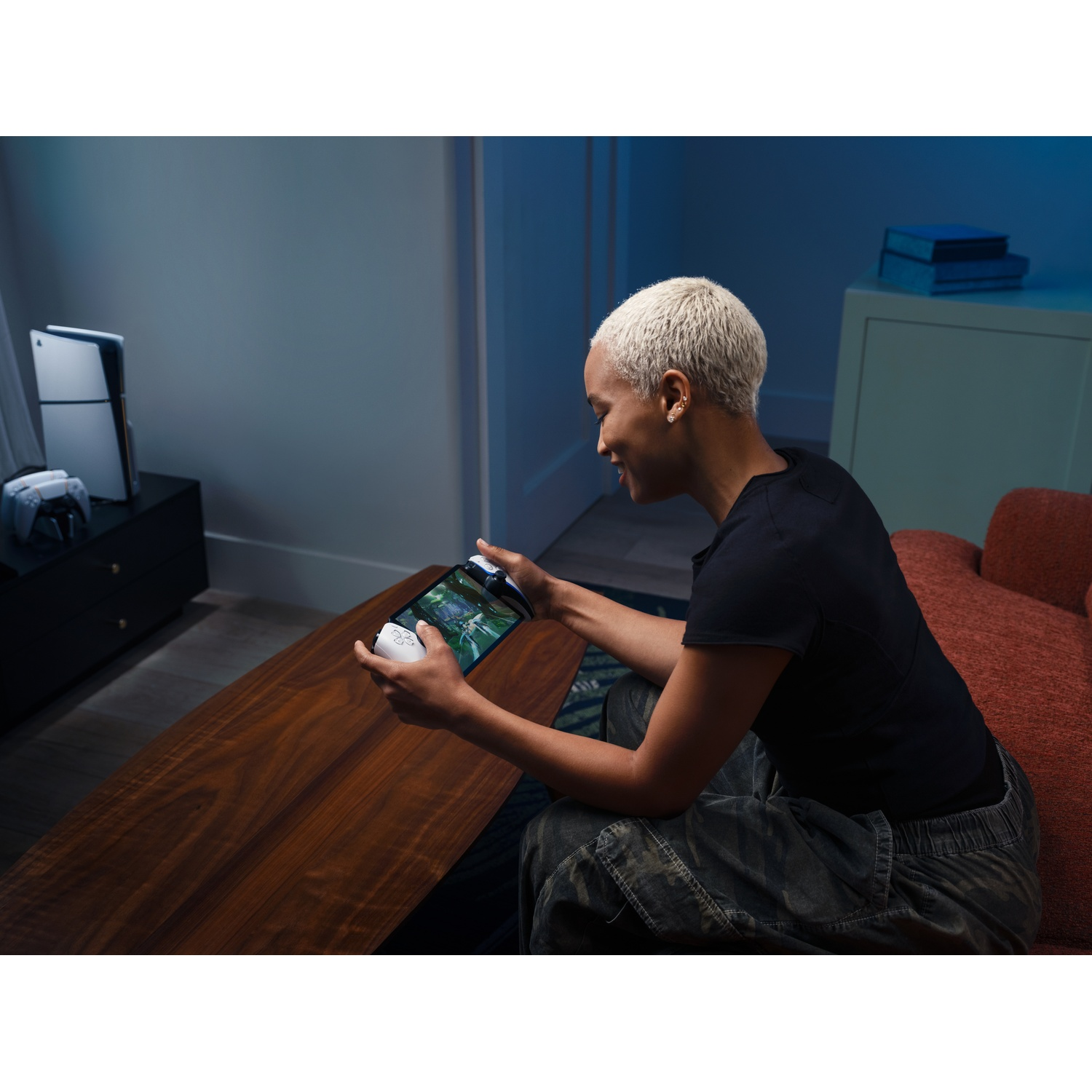 A person with short blonde hair is playing a handheld game console. In the background a PS5 and a sofa are visible.