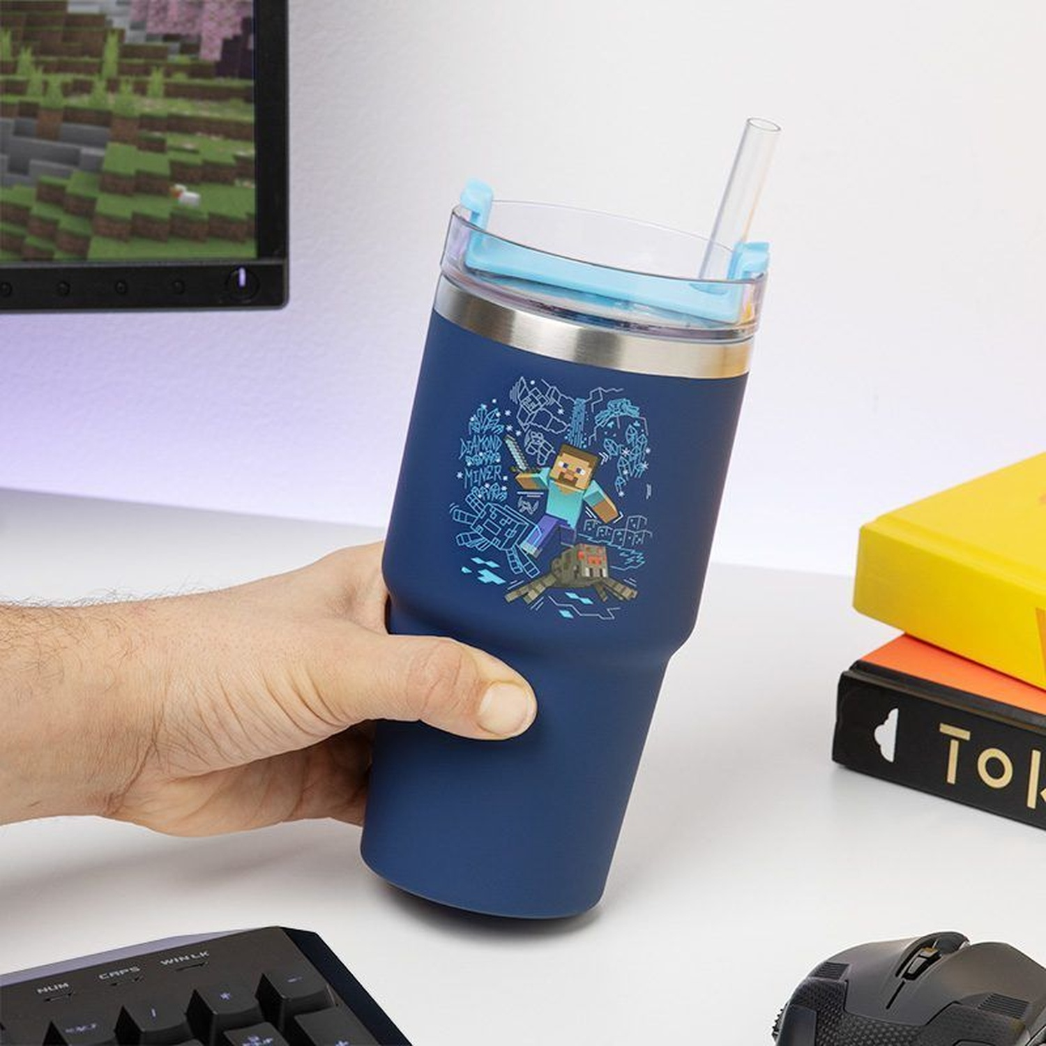 A hand holds a blue Minecraft tumbler with a straw. A keyboard, mouse, and books are on a white desk. A monitor displays a game.