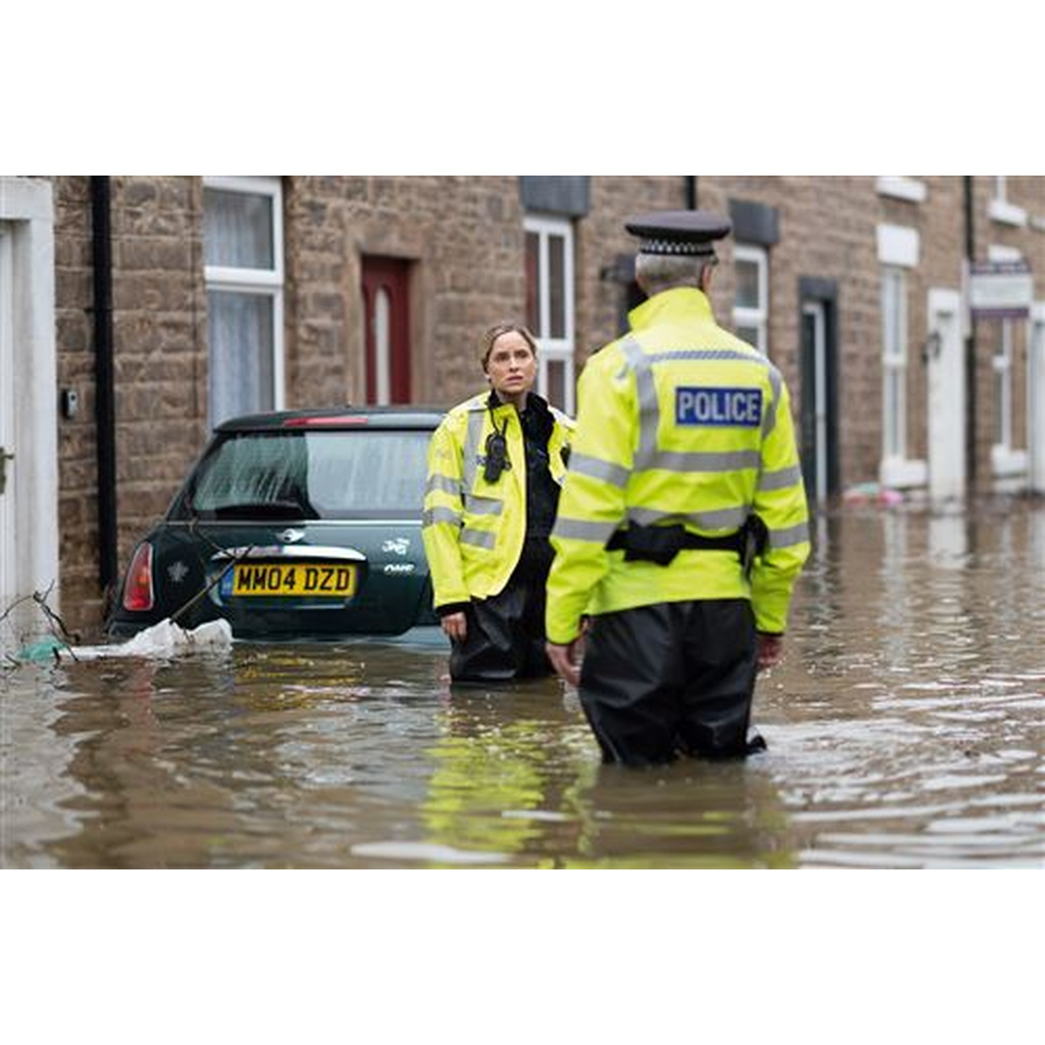 Two police officers stand in floodwater near a submerged car. Buildings are in the background, showing the impact of the flood.