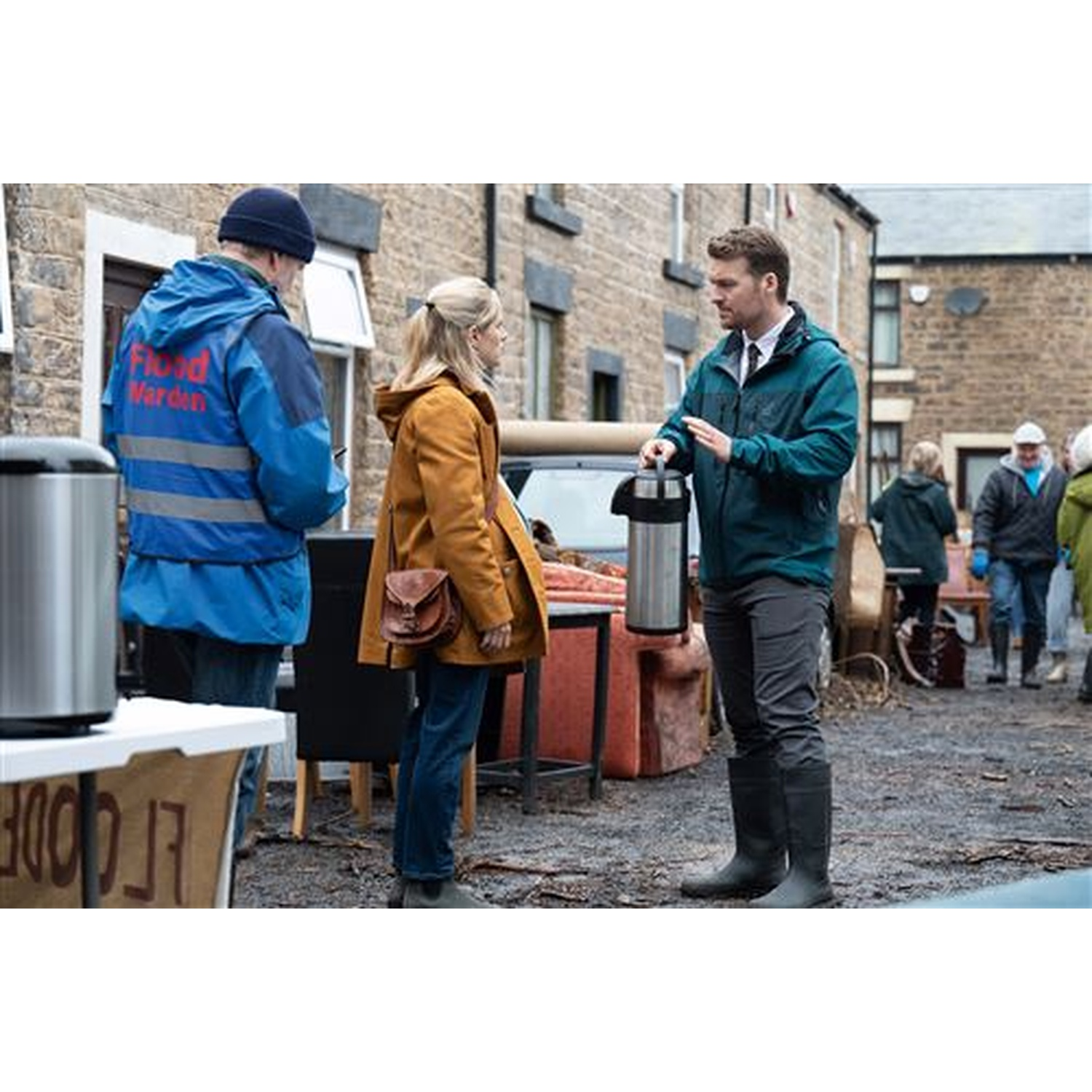 People in a street, one holding a thermos. Others are wearing jackets. A flood warden is present. Buildings are in the background.