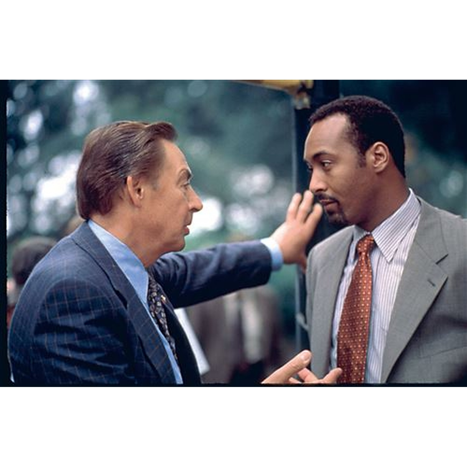 Two men in suits face each other, one touching the other's face. Green foliage in the background, suggesting an outdoor setting.