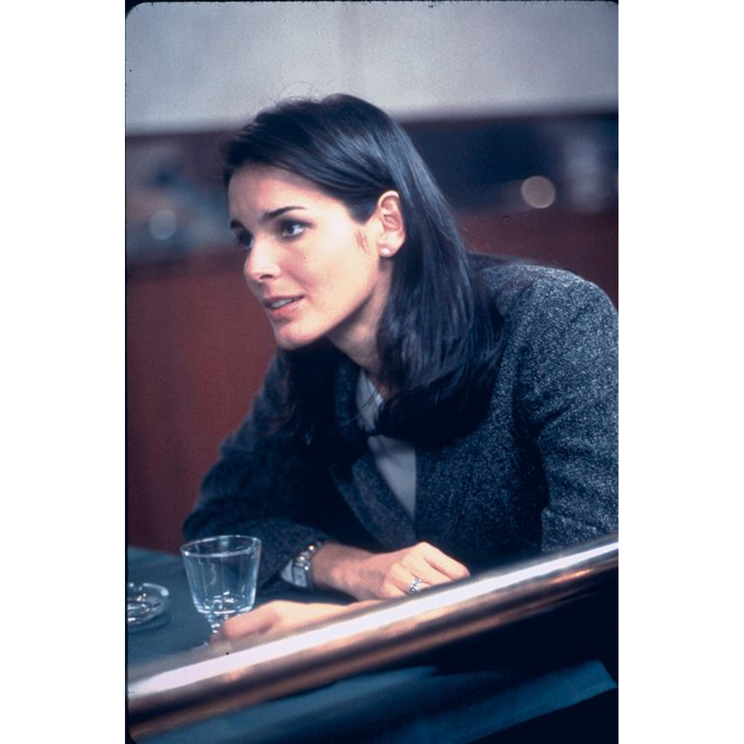 A woman with dark hair sits at a table. She wears a gray jacket and has a glass of water and an ashtray in front of her.