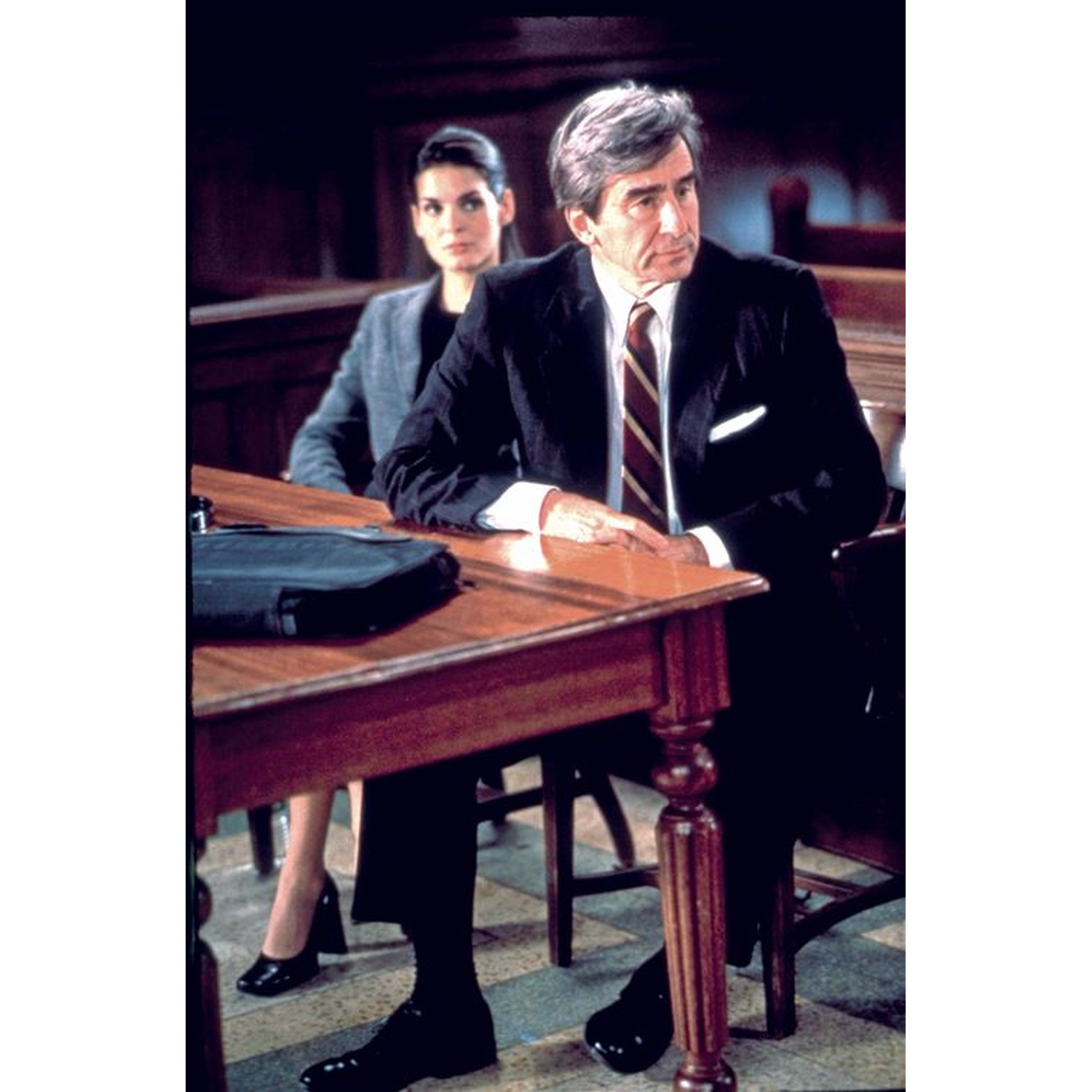 A man in a suit sits at a table. A woman sits behind him. There is a briefcase on the table.