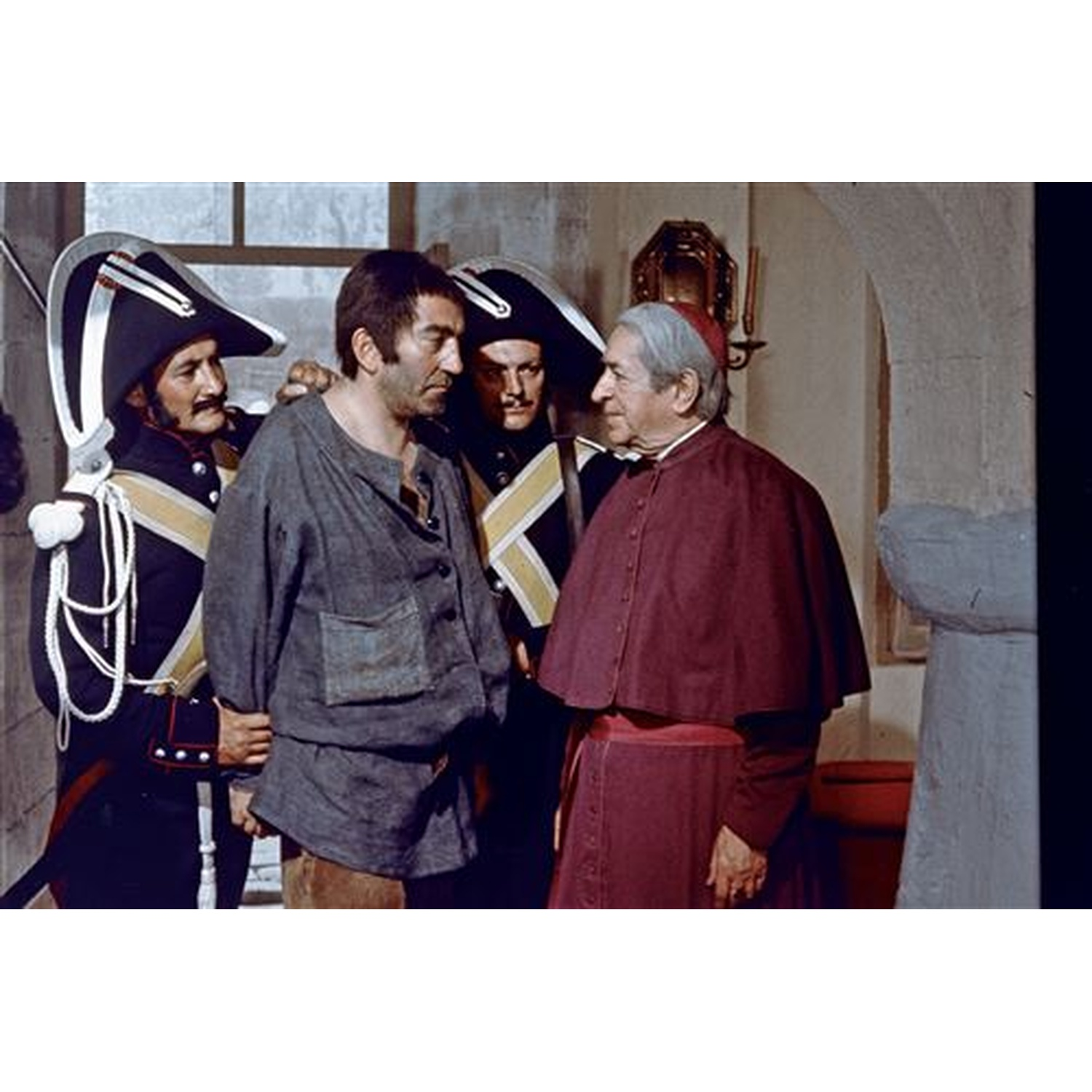 Un homme en gris est tenu par des gardes, face à un cardinal en robe rouge. Des soldats en chapeaux noirs montent la garde. La scène est à l'intérieur.