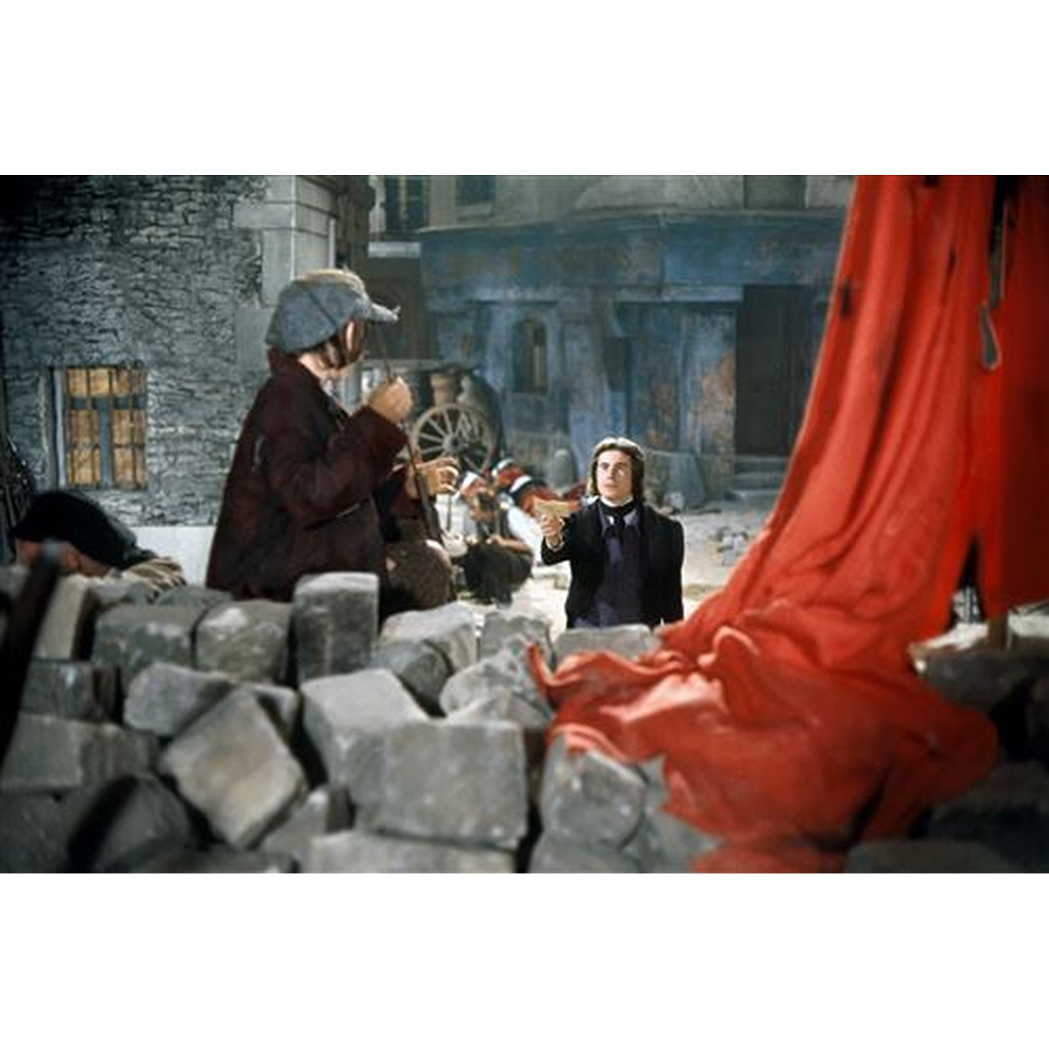 Deux hommes en tenue historique se tiennent près d'une barricade de pierres, avec un drap rouge. Des bâtiments et un chariot sont en arrière-plan.