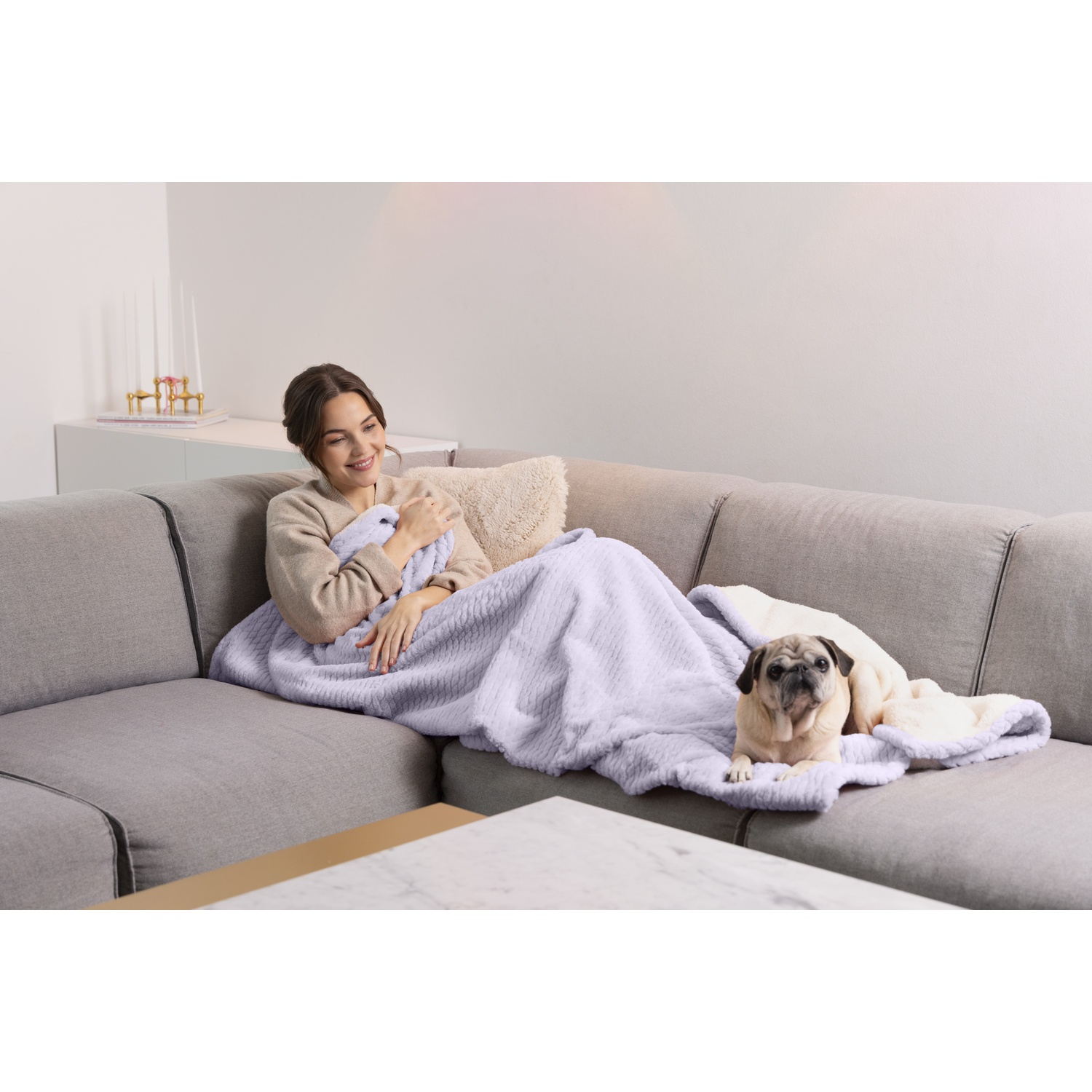 A woman relaxes on a sofa with a blanket and a pug dog. The dog is lying on the blanket.