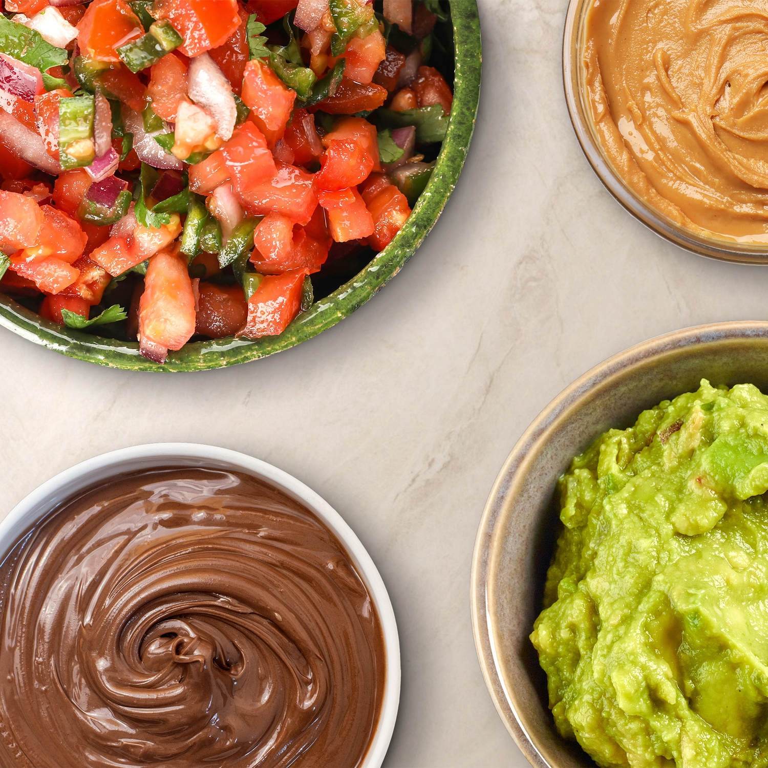 Overhead shot of four bowls: salsa, peanut butter, chocolate spread, and guacamole. All on a white marble surface.