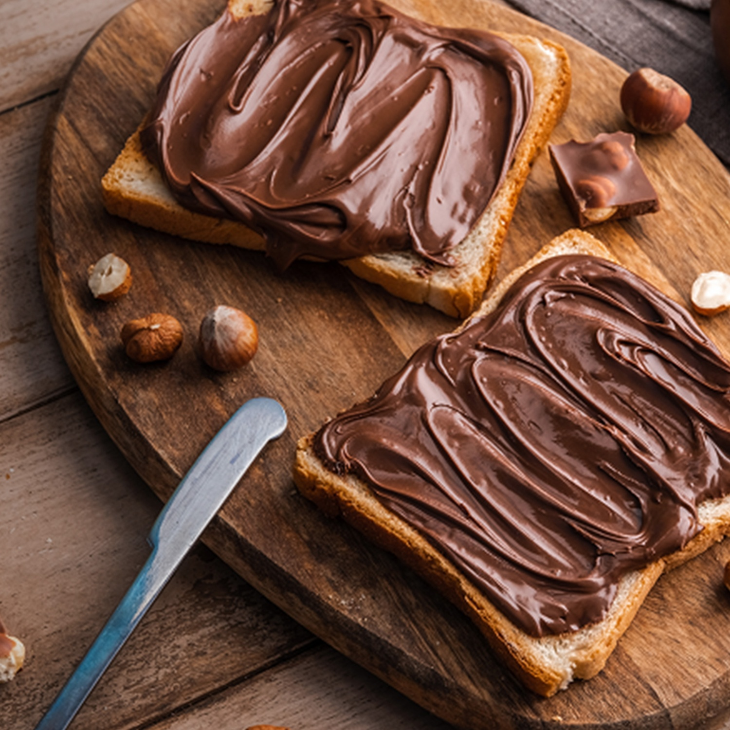 Two slices of toast spread with chocolate hazelnut spread, hazelnuts, and a knife on a wooden board.