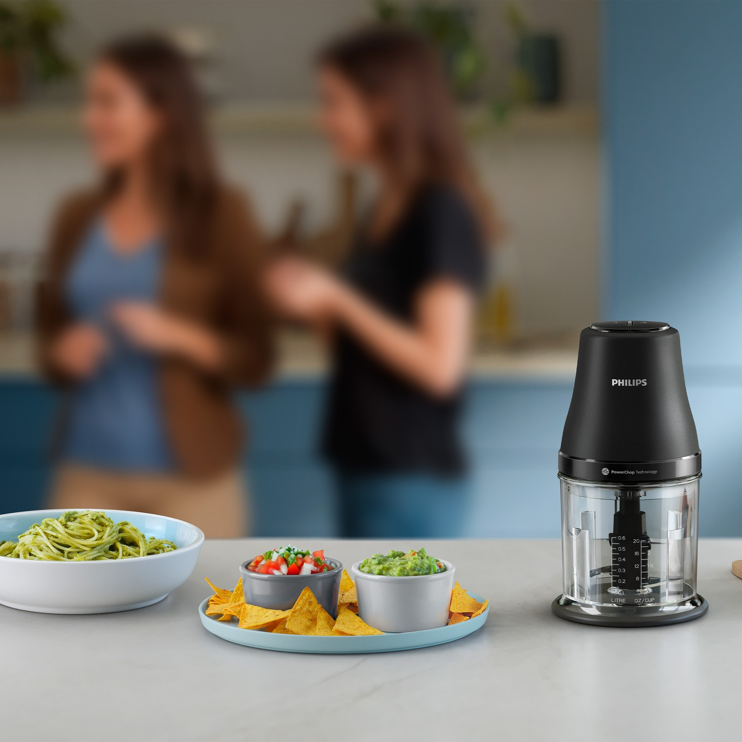 A table with pasta, salsa, guacamole, and a Philips food chopper. Two blurred women stand in the background of the kitchen scene.