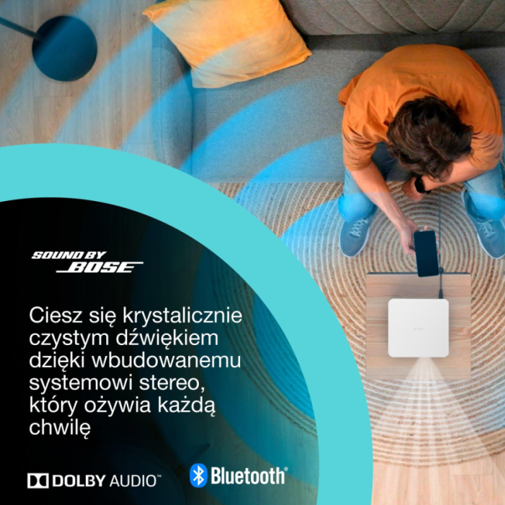 Osoba siedzi, używając telefonu podłączonego do białego głośnika na drewnianym stole. Widoczna jest sofa z żółtą poduszką.