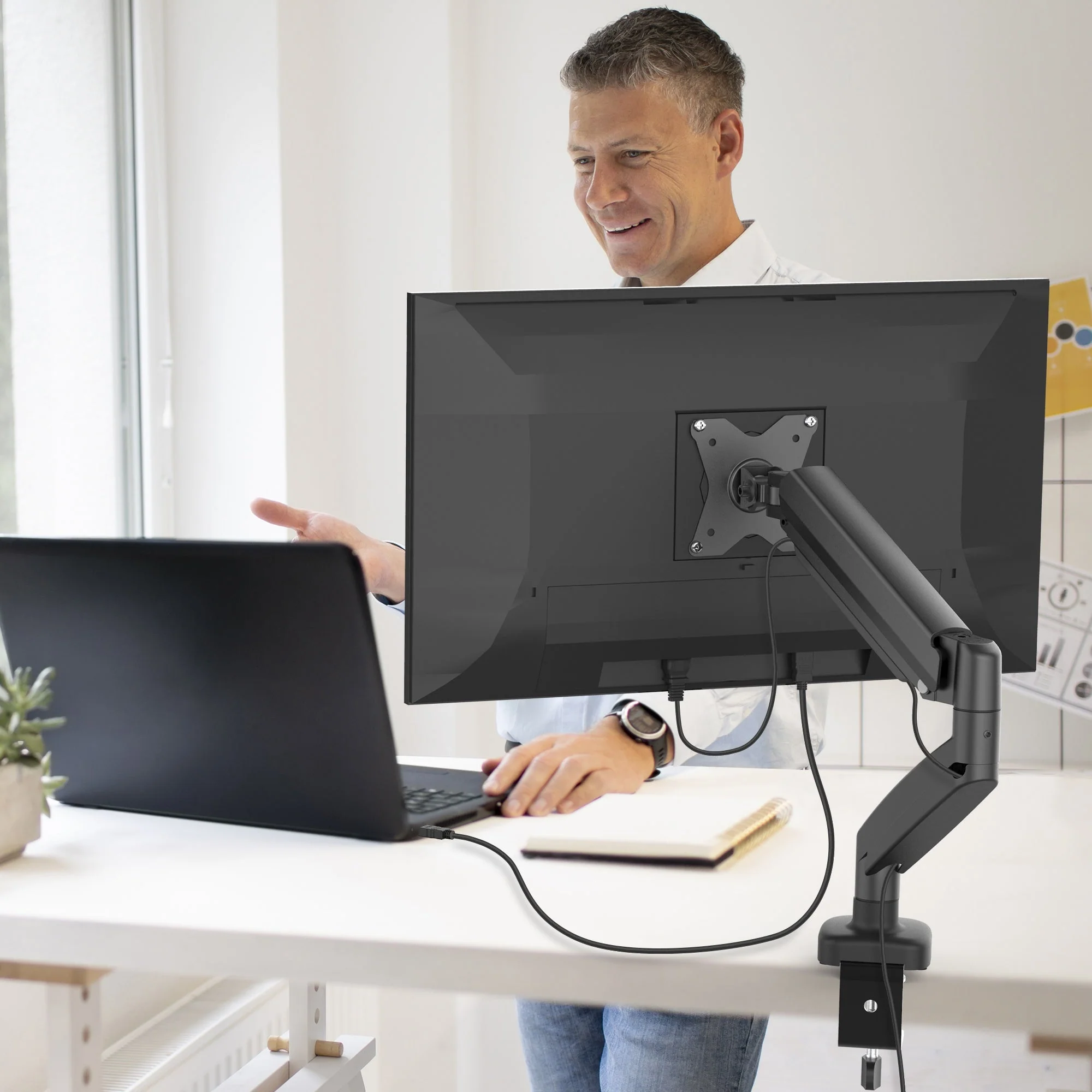 Férfi az asztalnál laptop-pal és monitorral. Kezével gesztikulál, mosolyog.