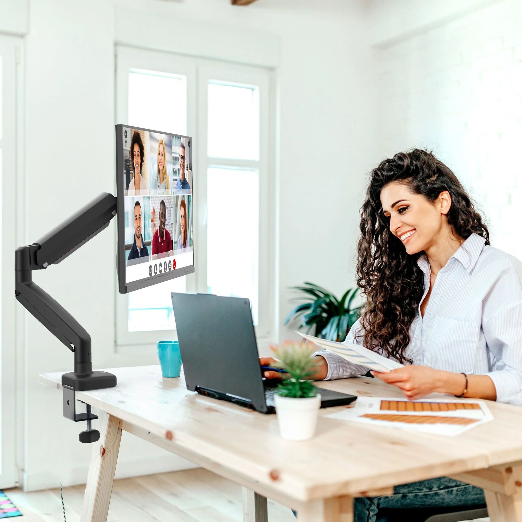 Nő videohívásban, laptopon dolgozik. A monitor videokonferenciát mutat. Asztalon növény, bögre, papírok. Fehér szoba.