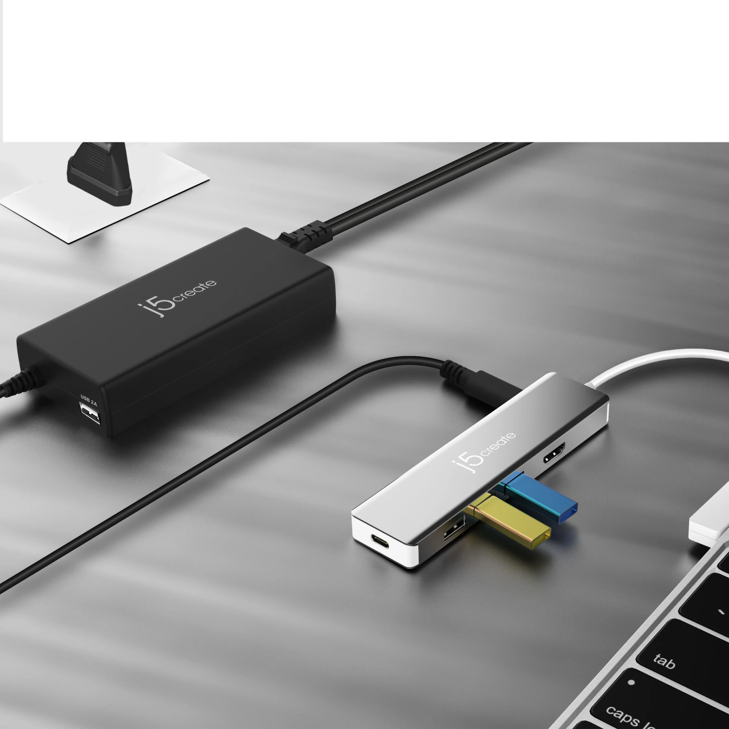 Egy fekete hálózati adapter és egy ezüst USB-elosztó csatlakoztatott kábelekkel. Egy laptop billentyűzete jobb alsó sarokban.