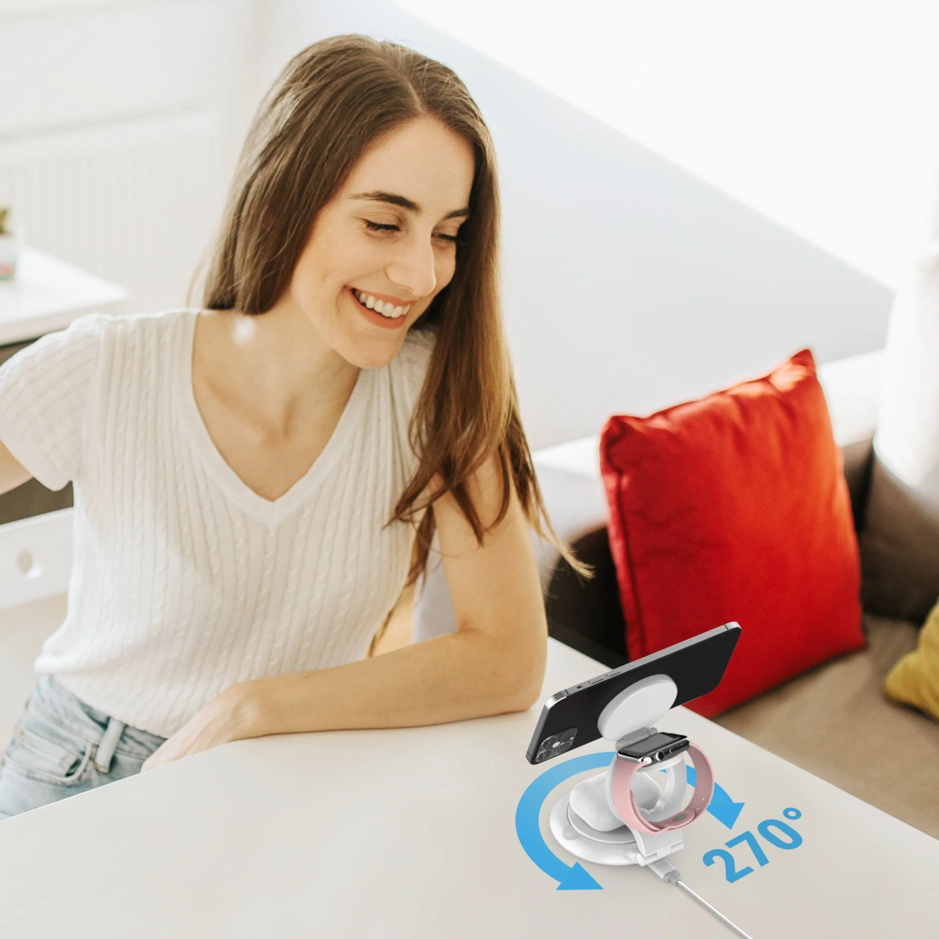 Egy nő mosolyog egy telefon- és óratöltőn egy asztalon. Vörös párna és kanapé a háttérben. 270 fokos nyíl.