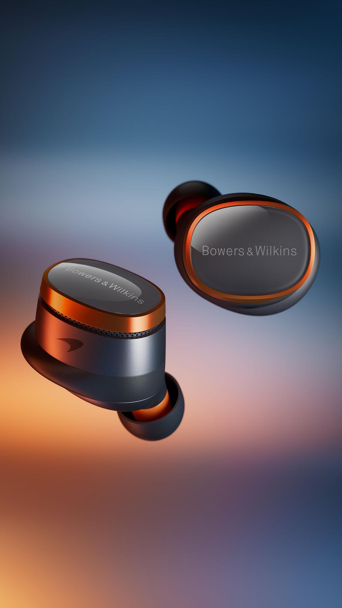 Zwei Bowers & Wilkins Ohrhörer, dunkelgrau mit orangefarbenen Akzenten, schweben vor einem blauen und orangefarbenen Hintergrund.