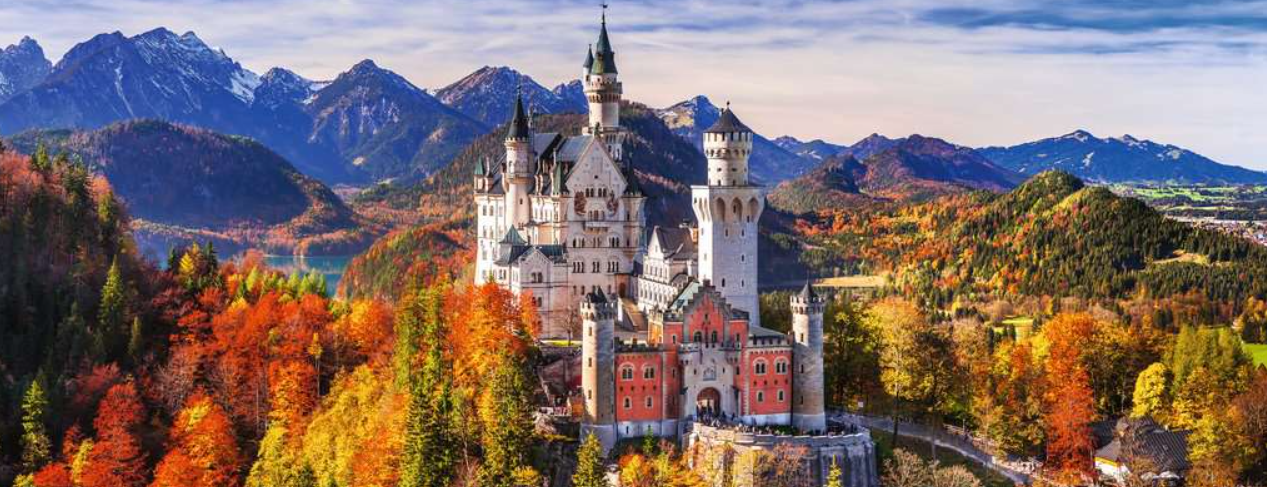 Schloss Neuschwanstein im Herbst. Berge im Hintergrund, bunte Bäume und ein See. Klarer Himmel.