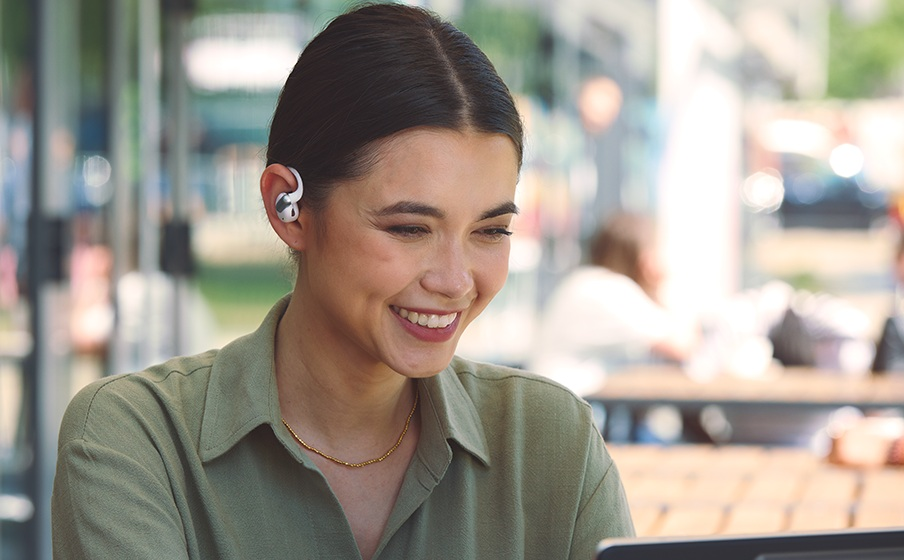 Kobieta uśmiecha się, używając laptopa na zewnątrz. Ma słuchawki i złoty naszyjnik. Inni ludzie są rozmyci w tle.
