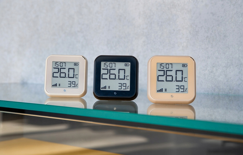 Trzy cyfrowe termometry wyświetlające czas, temperaturę (26,0°C) i wilgotność (39%). Każdy ma inną kolorową ramkę: beżową, czarną i beżową.
