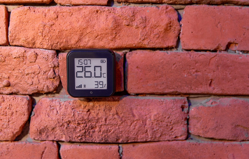 Czarne urządzenie cyfrowe wyświetla godzinę, temperaturę (26,0°C) i wilgotność (39%) na czerwonej ceglanej ścianie.