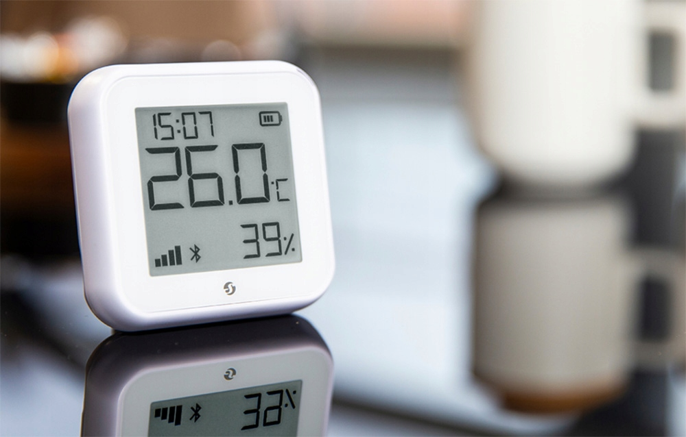 Biały wyświetlacz cyfrowy pokazuje czas, temperaturę (26,0°C) i wilgotność (39%). Odbija się na powierzchni. Kubek jest rozmyty w tle.