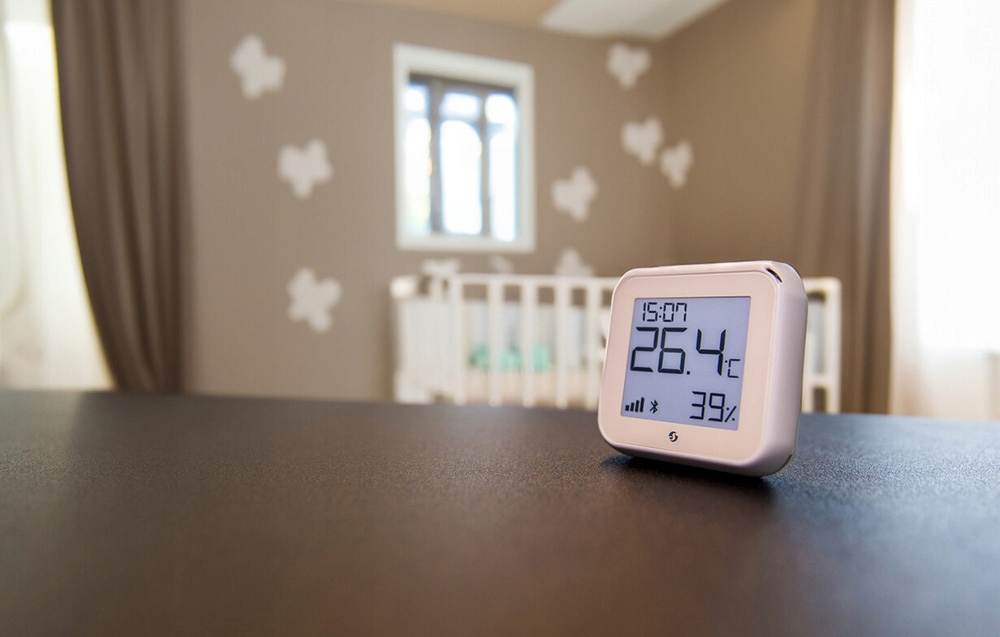Cyfrowy wyświetlacz pokazuje czas, temperaturę i wilgotność. W tle widoczne jest łóżeczko, okno i dekoracyjne naklejki ścienne.